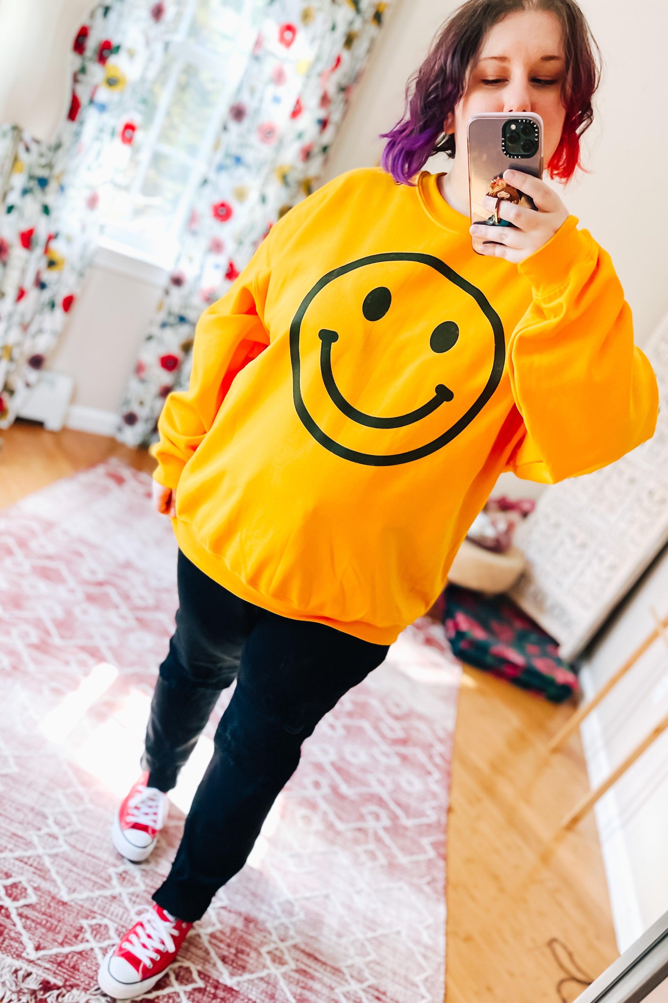 Woman wearing bright yellow smiley face sweatshirt with black pants and red shoes taking mirror selfie indoors