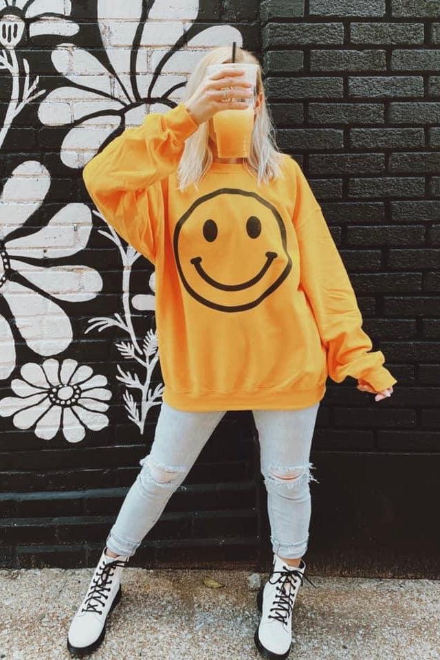Woman wearing oversized yellow sweatshirt with large black smiley face graphic holding matching yellow drink