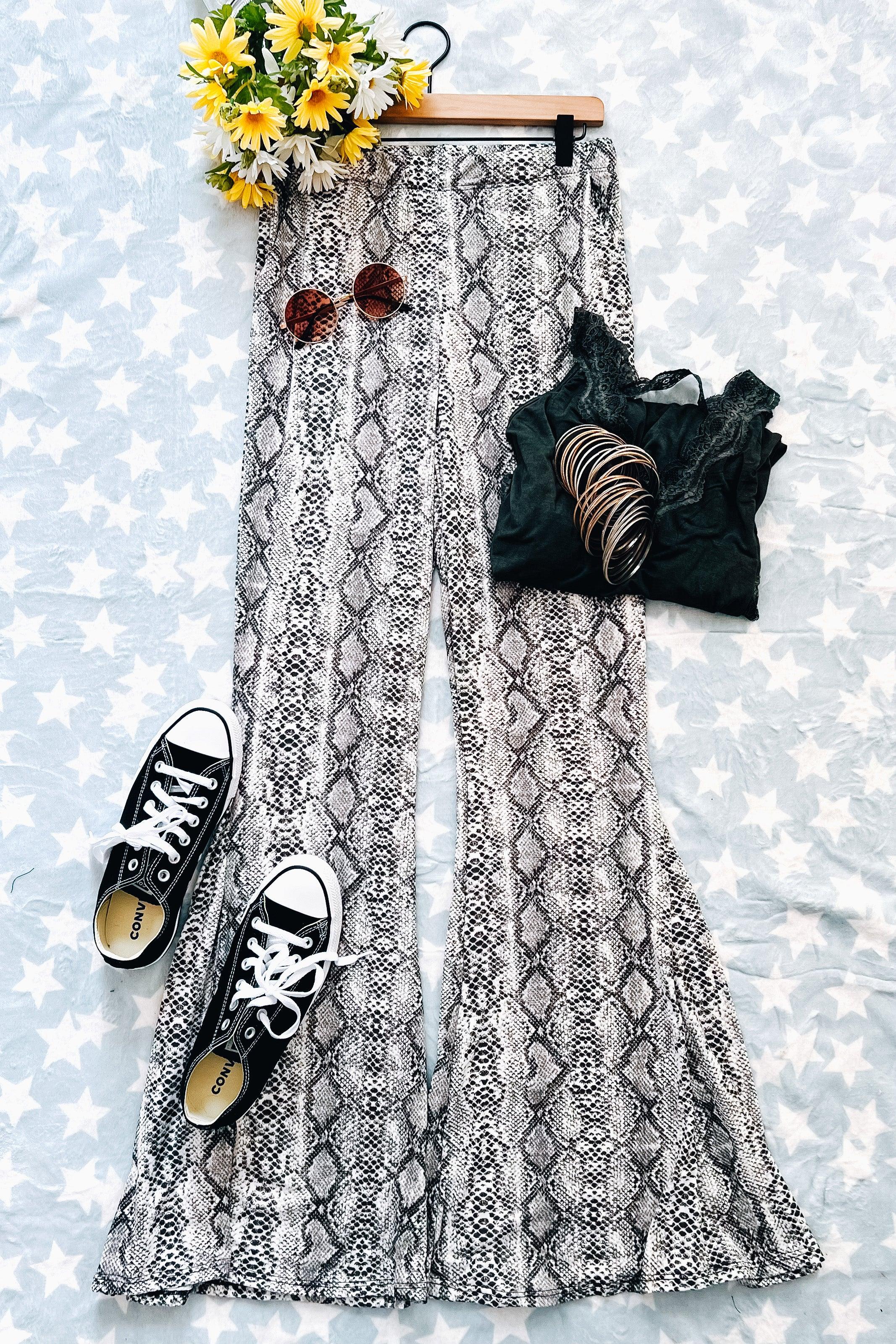 Grey snake print flare pants laid flat with black lace tank top, black sneakers, sunglasses, bangles, and yellow flowers