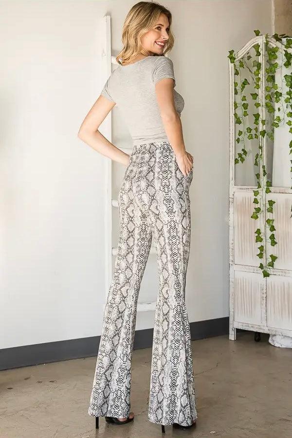 Smiling woman wearing grey snake print flare pants paired with fitted grey top standing near ivy-decorated screen