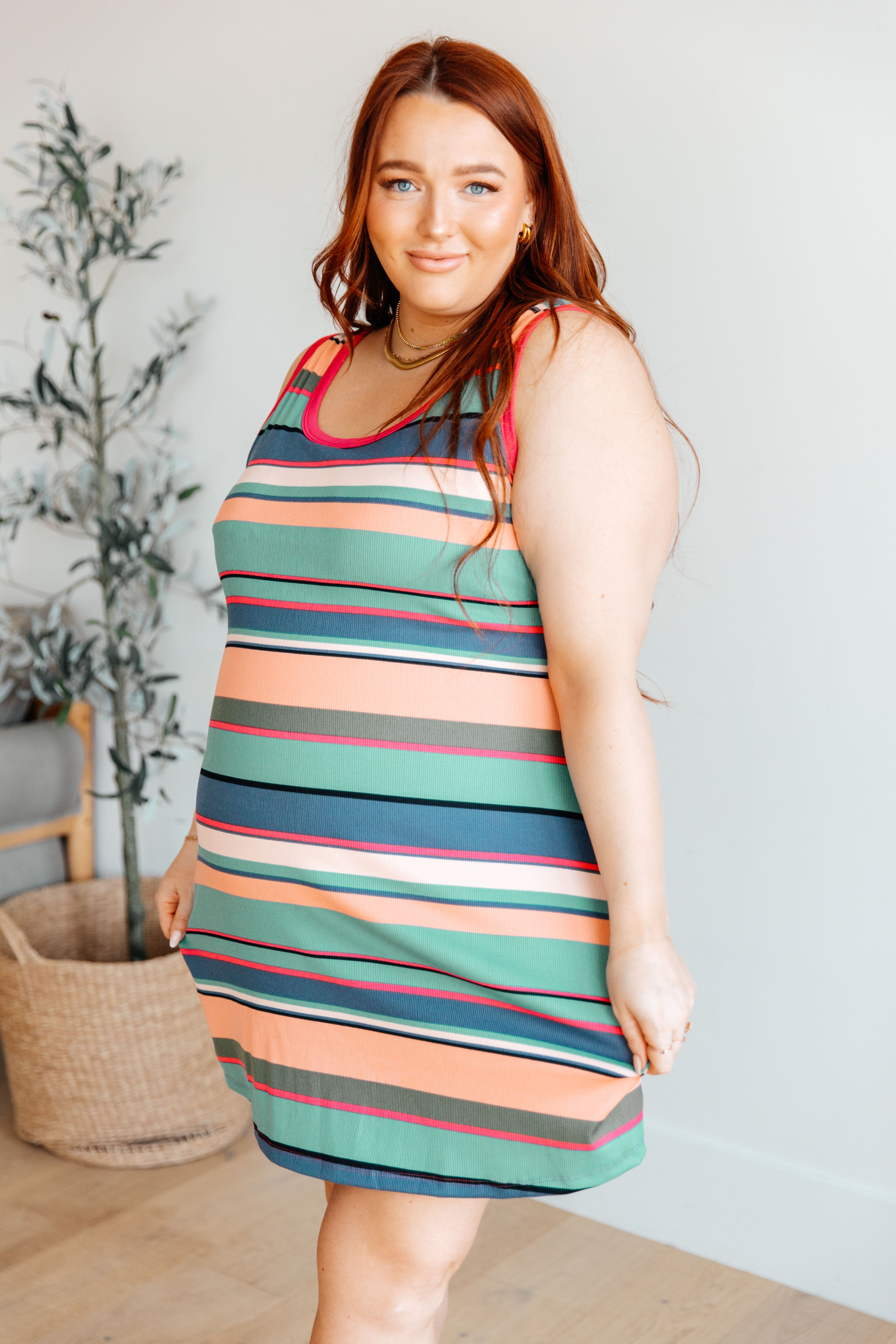 Curvy woman posing in a colorful striped tank dress with green, peach, navy, and red tones in a bright room