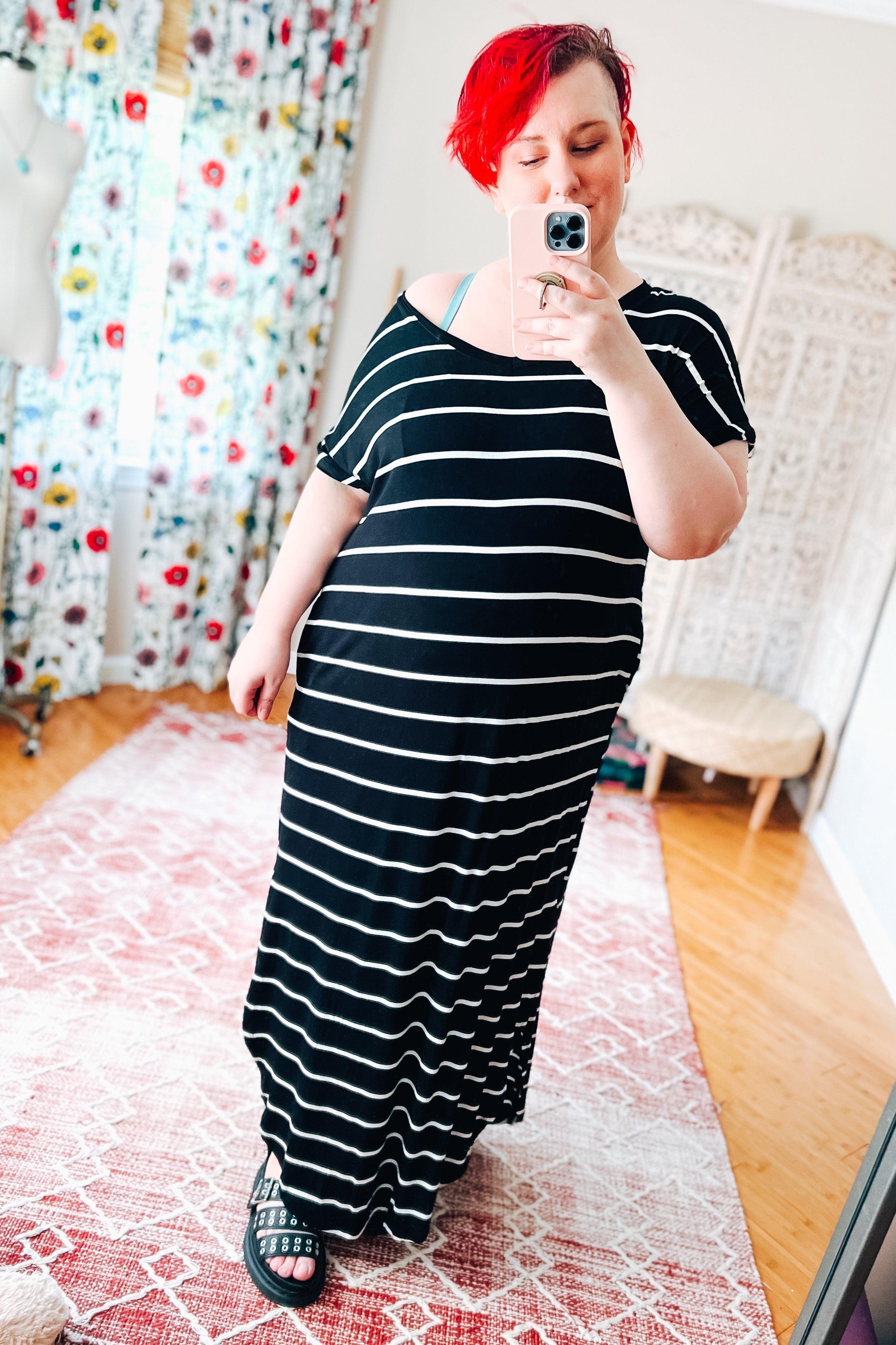 Woman with red hair wearing a black striped maxi dress taking a mirror selfie in a bright room with floral curtains