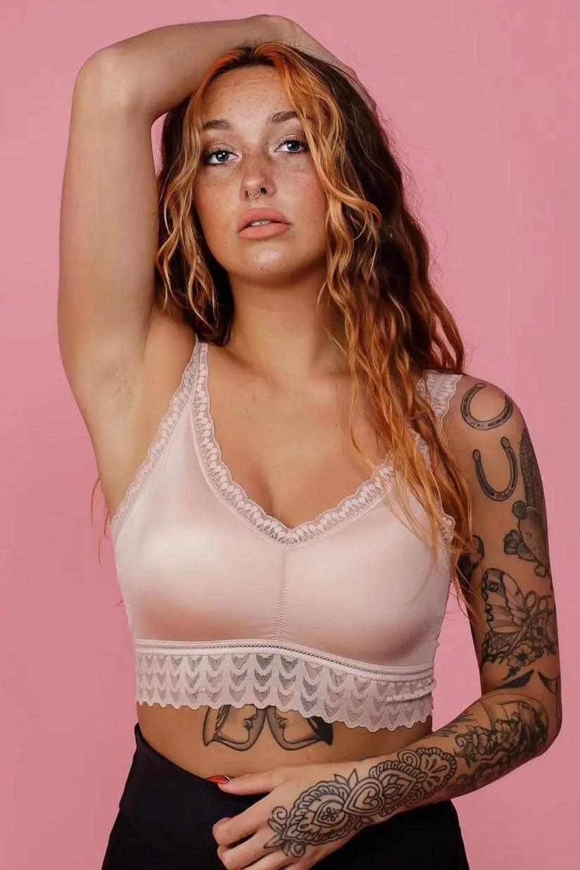 Woman with tattoos wearing a soft pink lace-trim bralette posing with one arm raised against pink background