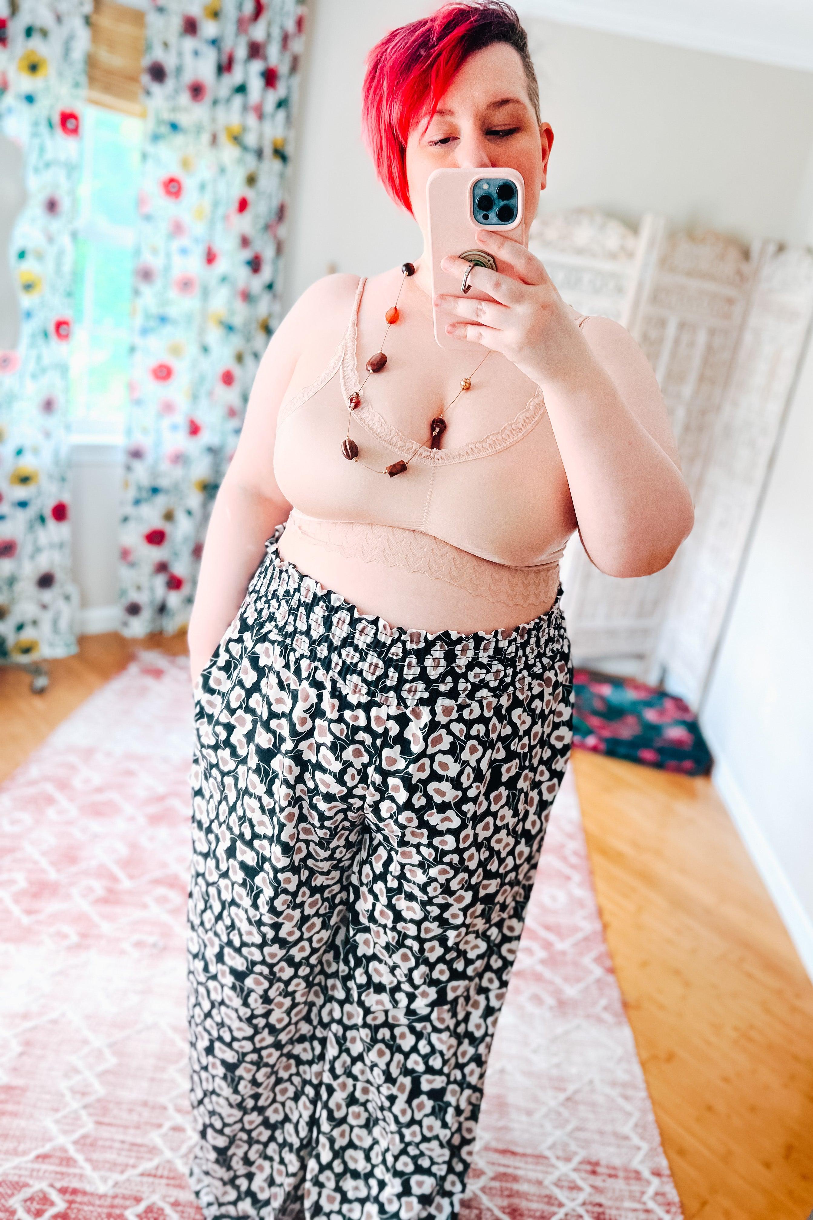 Plus-size woman wearing a soft pink bralette and floral wide-leg pants taking a mirror selfie indoors