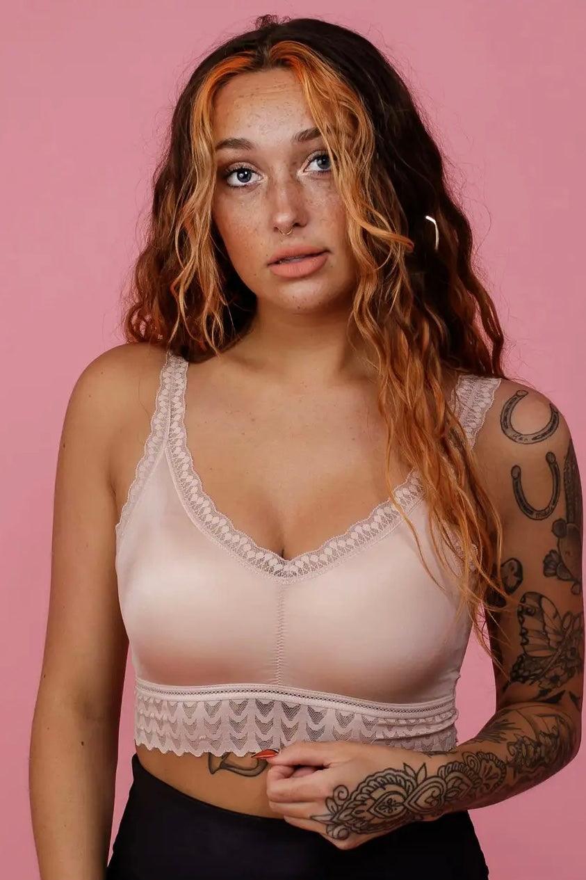 Woman with long wavy hair wearing a pink lace-trimmed bralette against a solid pink background