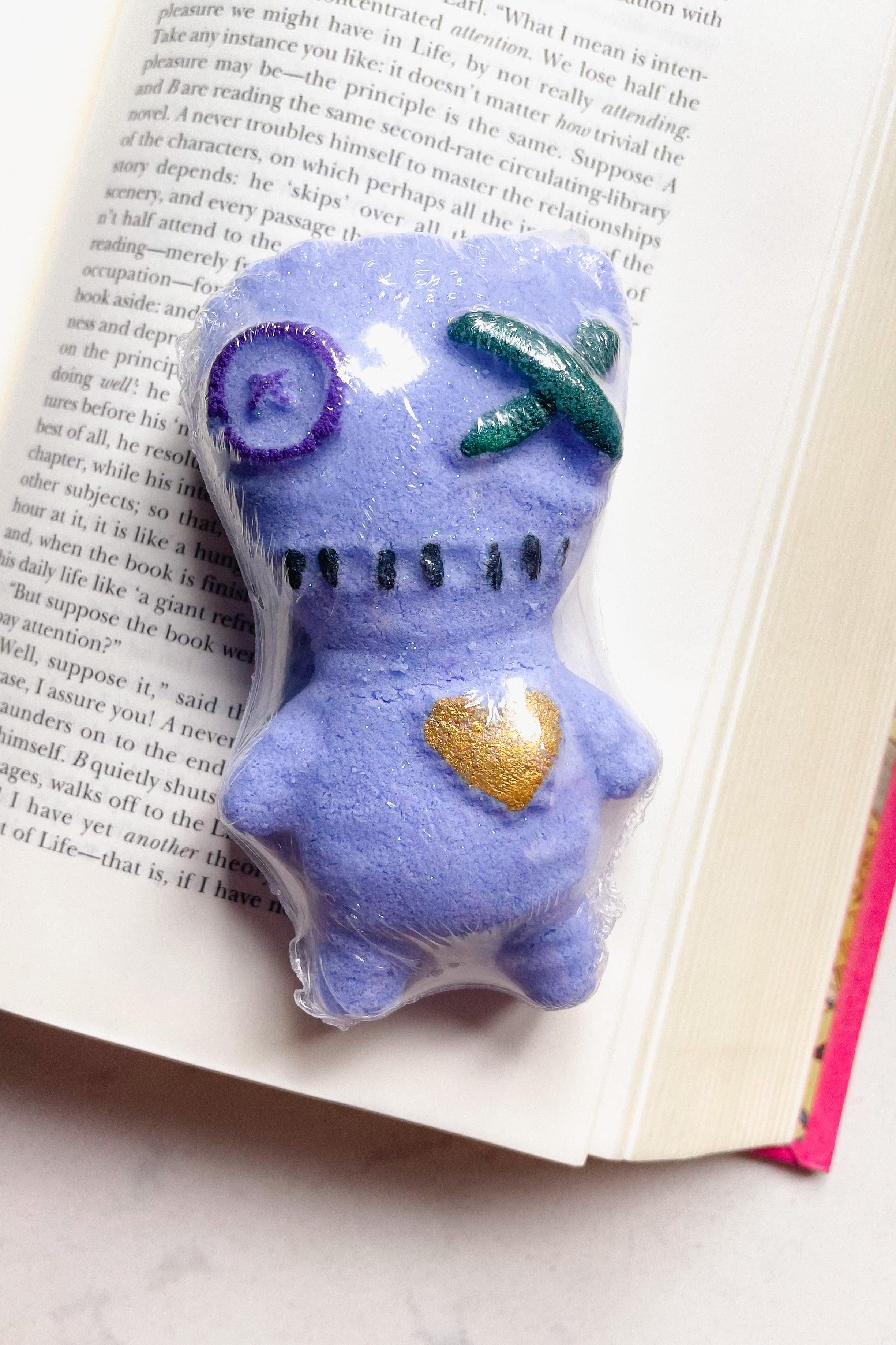 Purple voodoo doll shaped bath bomb with stitched mouth and decorative heart lying on open book pages