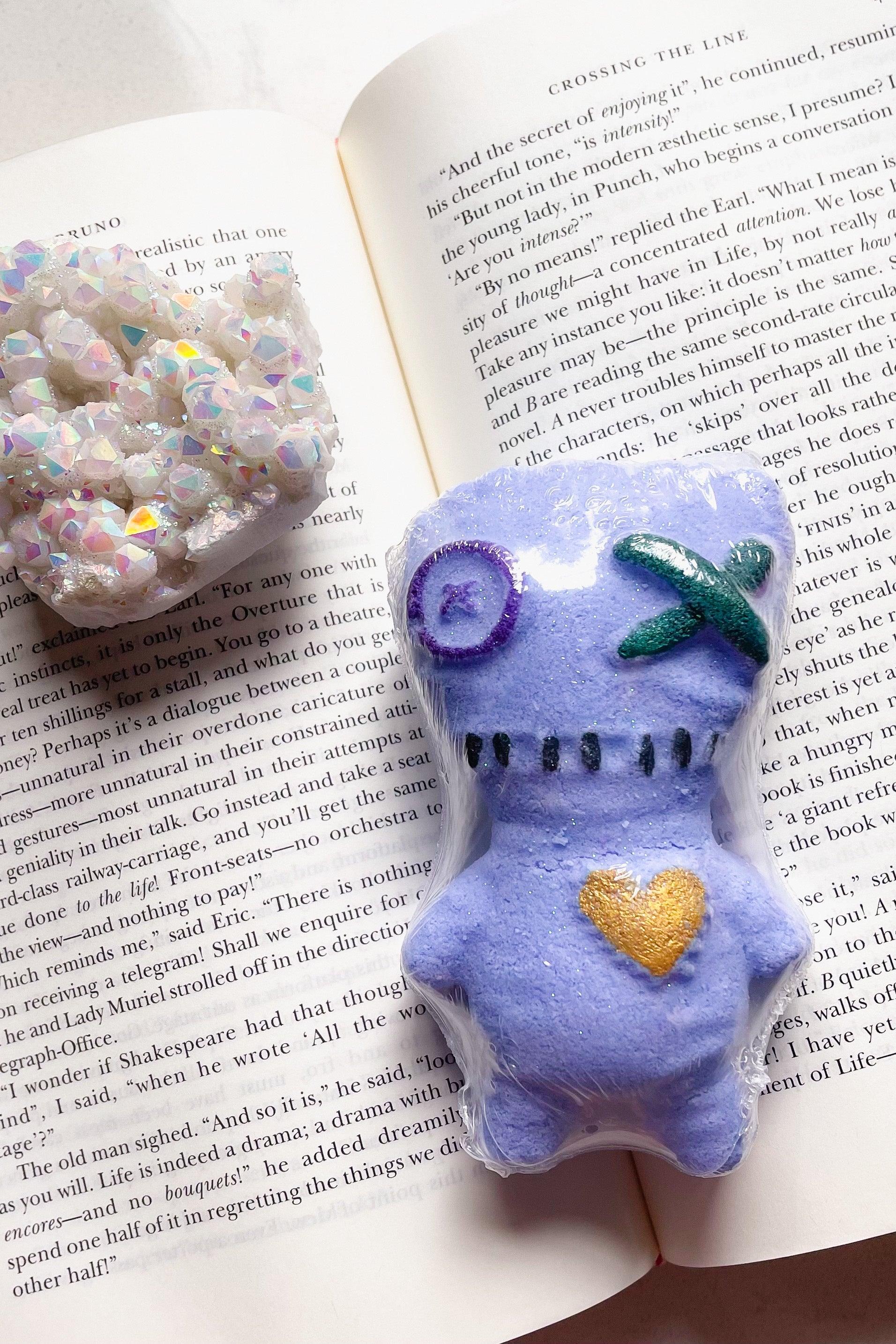 Purple voodoo doll-shaped bath bomb with stitched details and gold heart next to iridescent crystal cluster on book pages