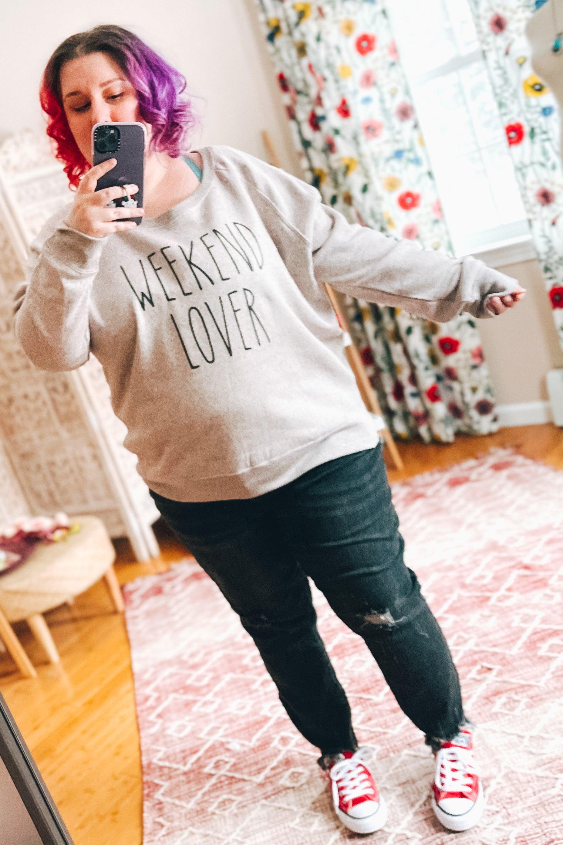 Woman with colorful hair wearing light gray relaxed pullover with "Weekend Lover" text paired with black ripped jeans indoors