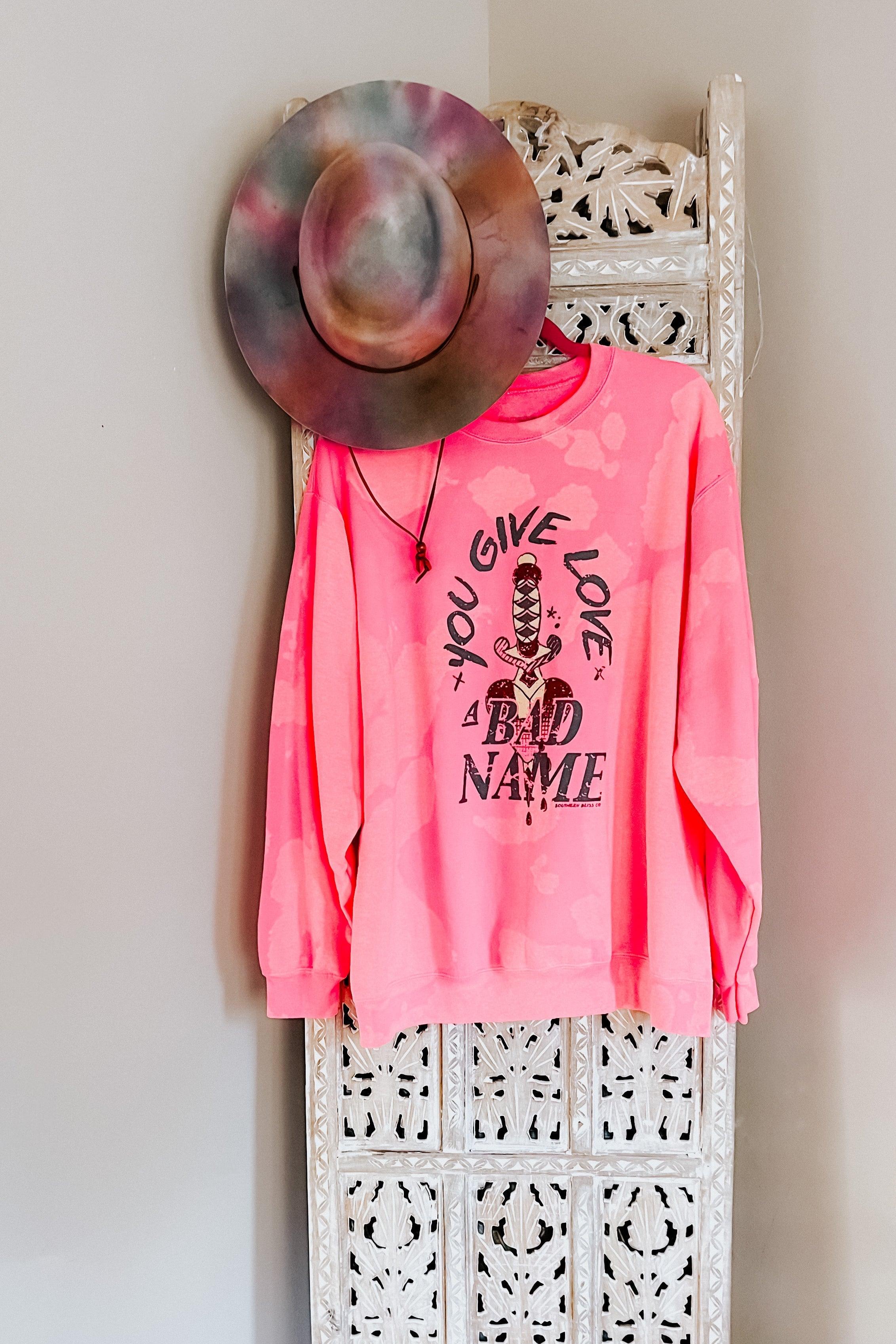 Pink bleached pullover with bold black sword graphic and text hanging next to a colorful tie-dye hat on carved wooden panel