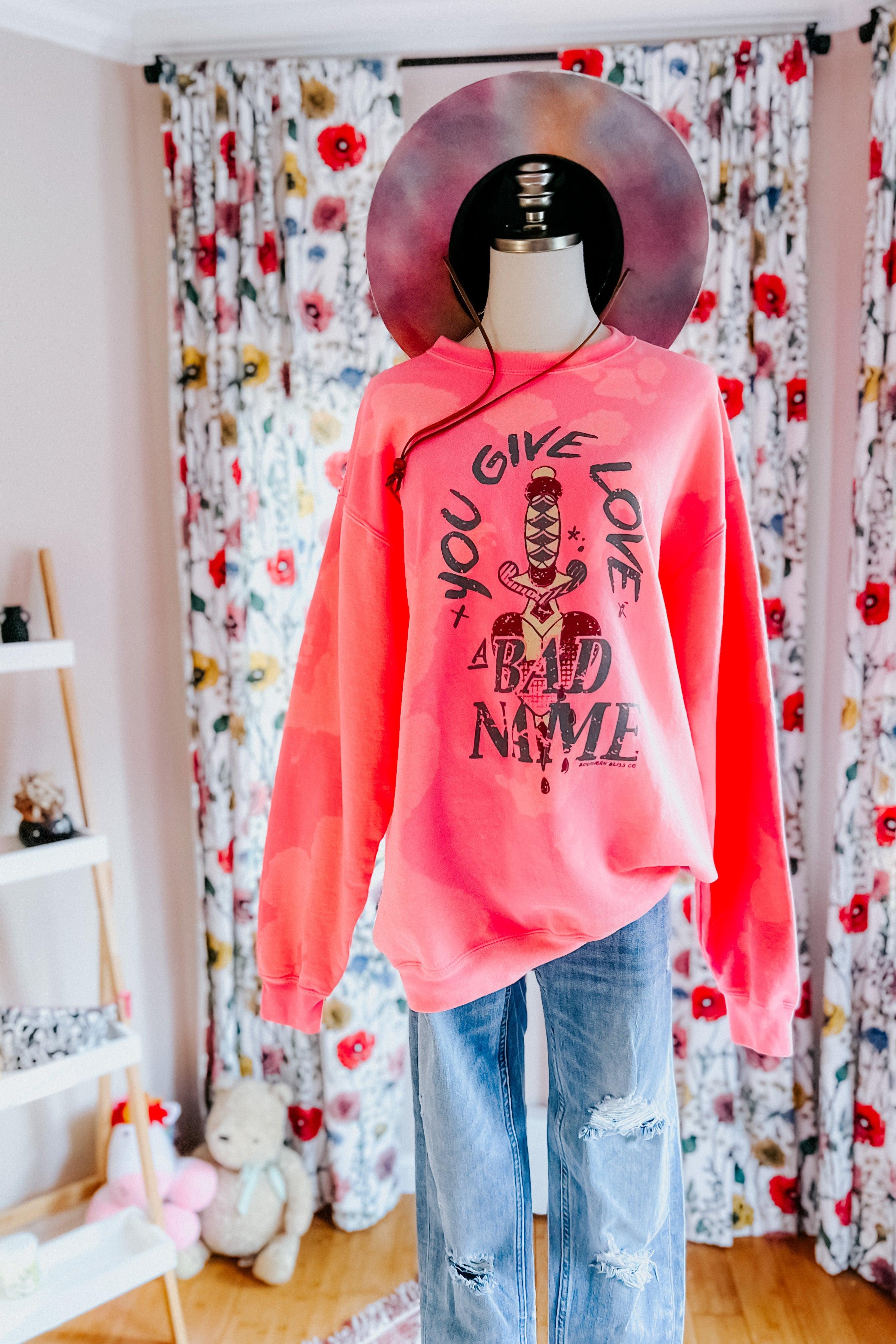 Mannequin dressed in pink bleached pullover with graphic lettering paired with ripped light blue jeans and hat indoors