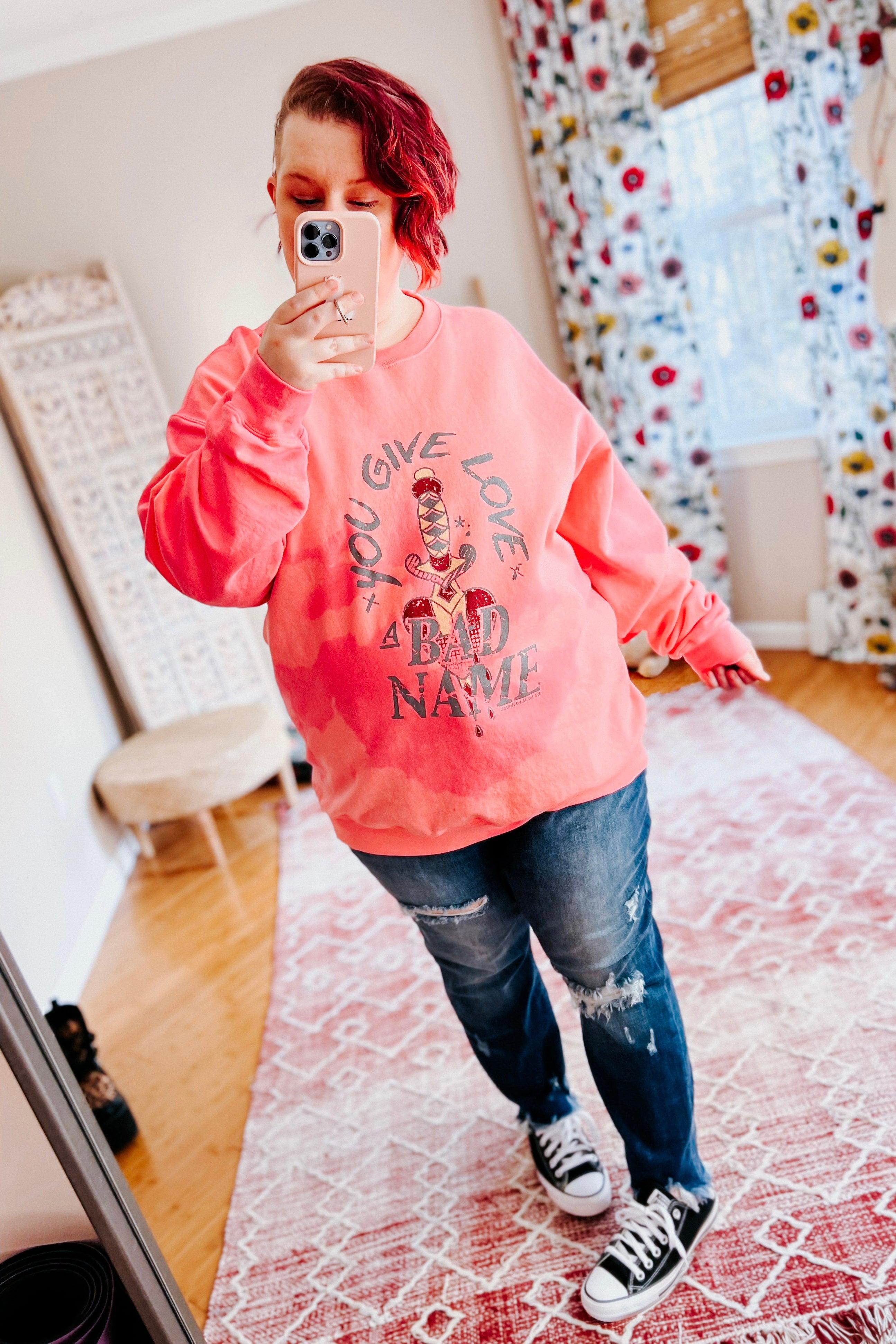 Woman wearing a bright pink bleached pullover with graphic text paired with ripped jeans and sneakers indoors