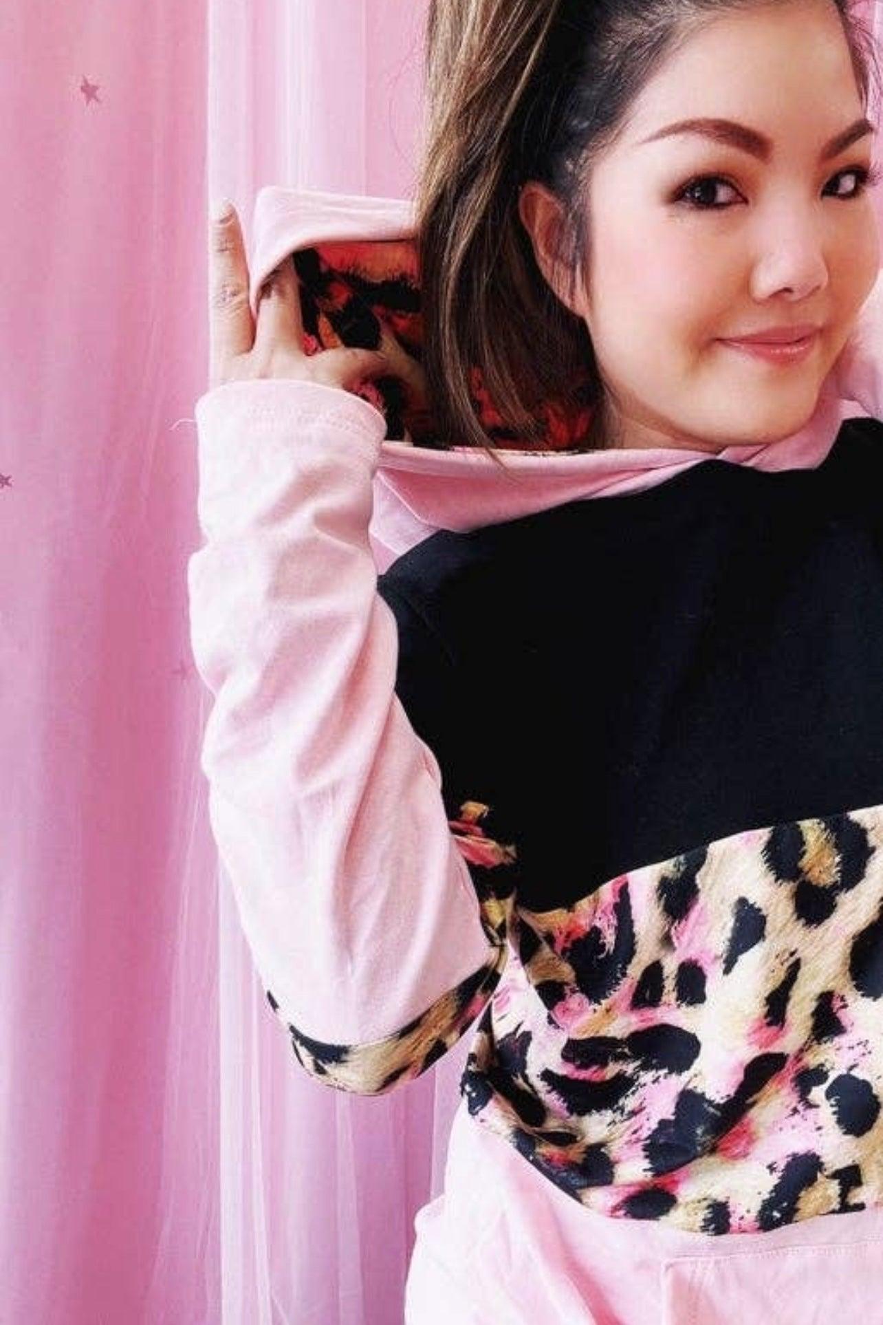 Woman wearing black and pink leopard print hoodie with light pink sleeves posing indoors against pink backdrop
