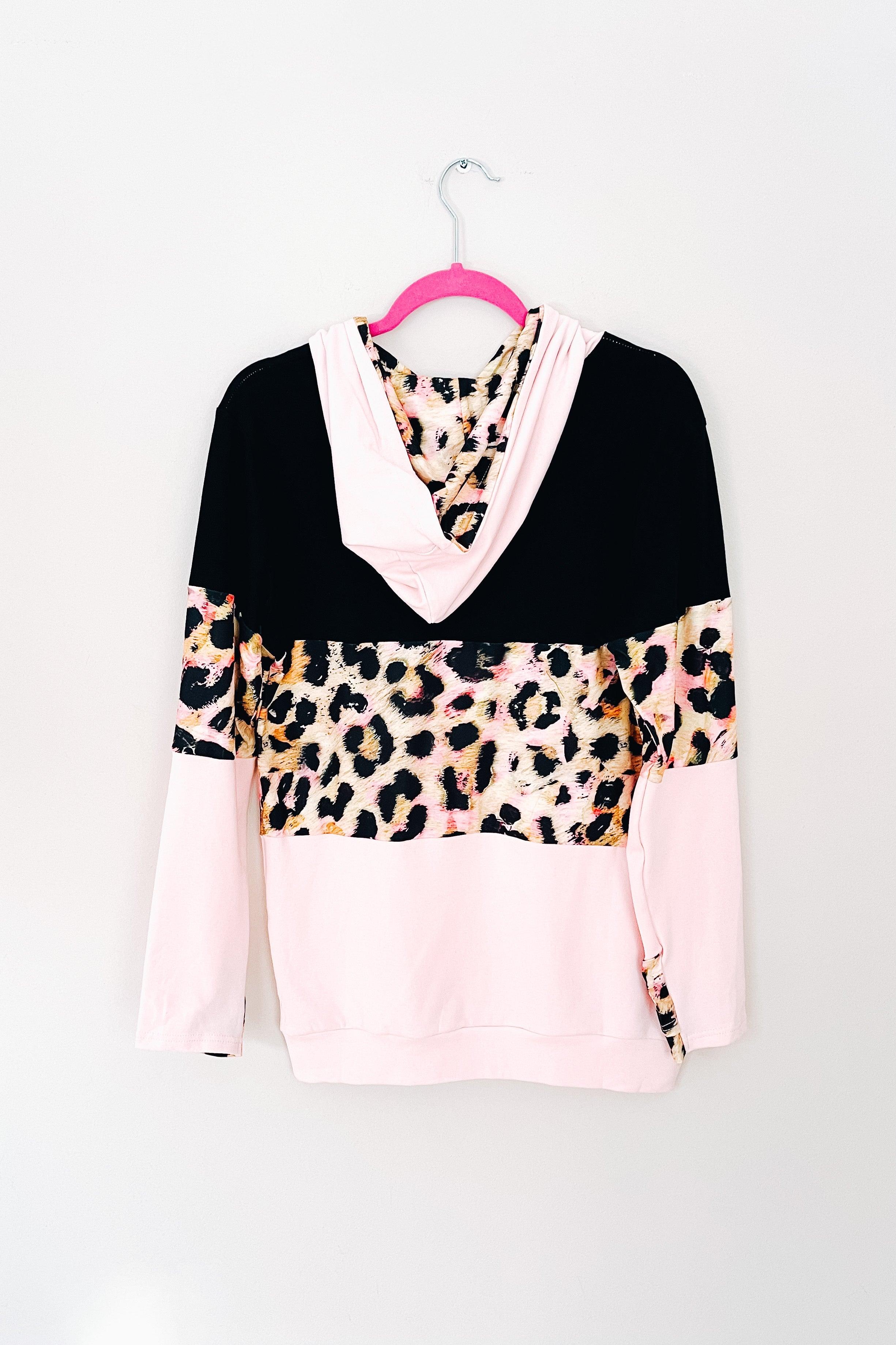 Black and pink hoodie with leopard print panel and light pink sleeves hanging on bright pink hanger against white wall