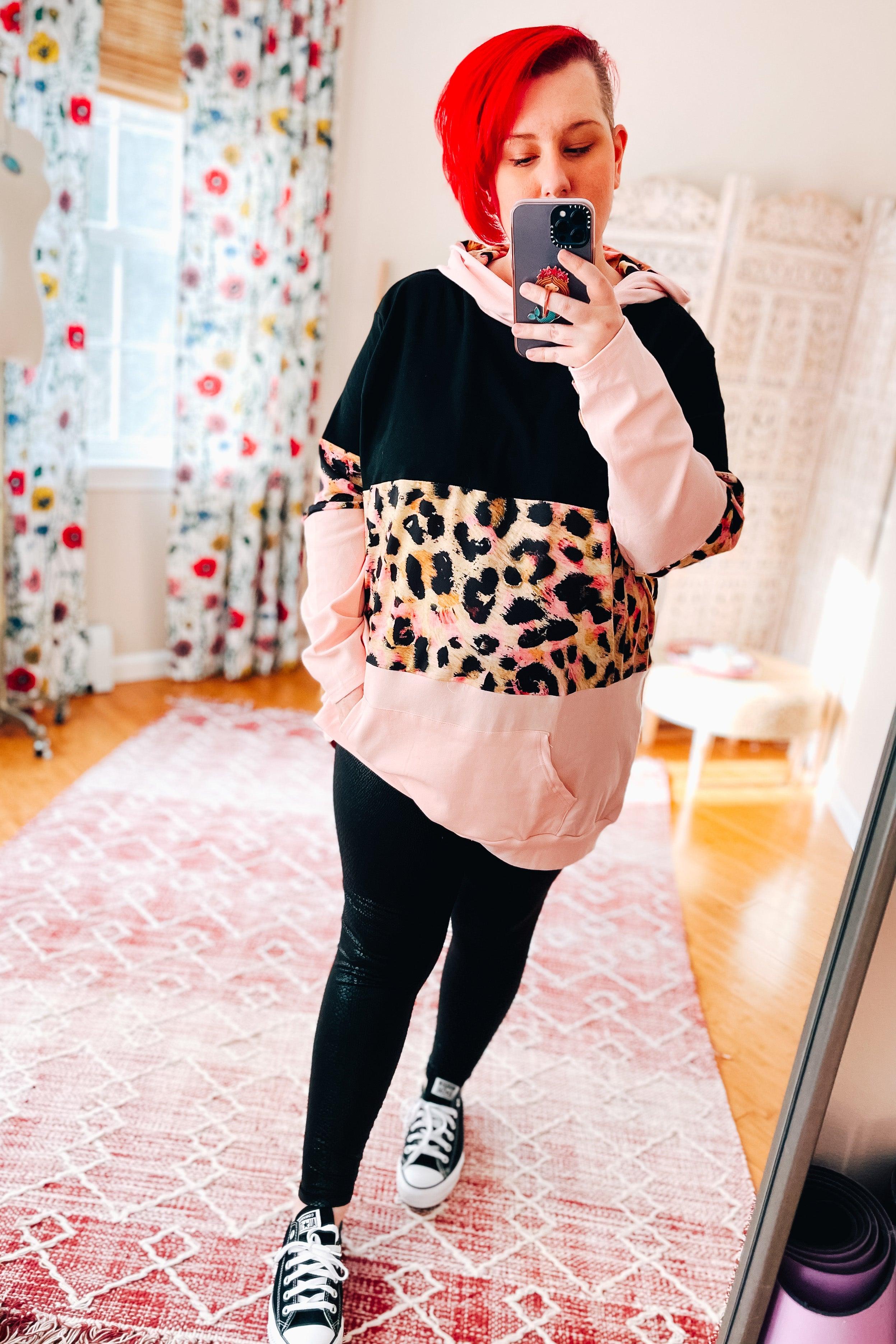Woman with bright red hair wearing a black and pink hoodie with leopard print panel paired with black leggings and sneakers