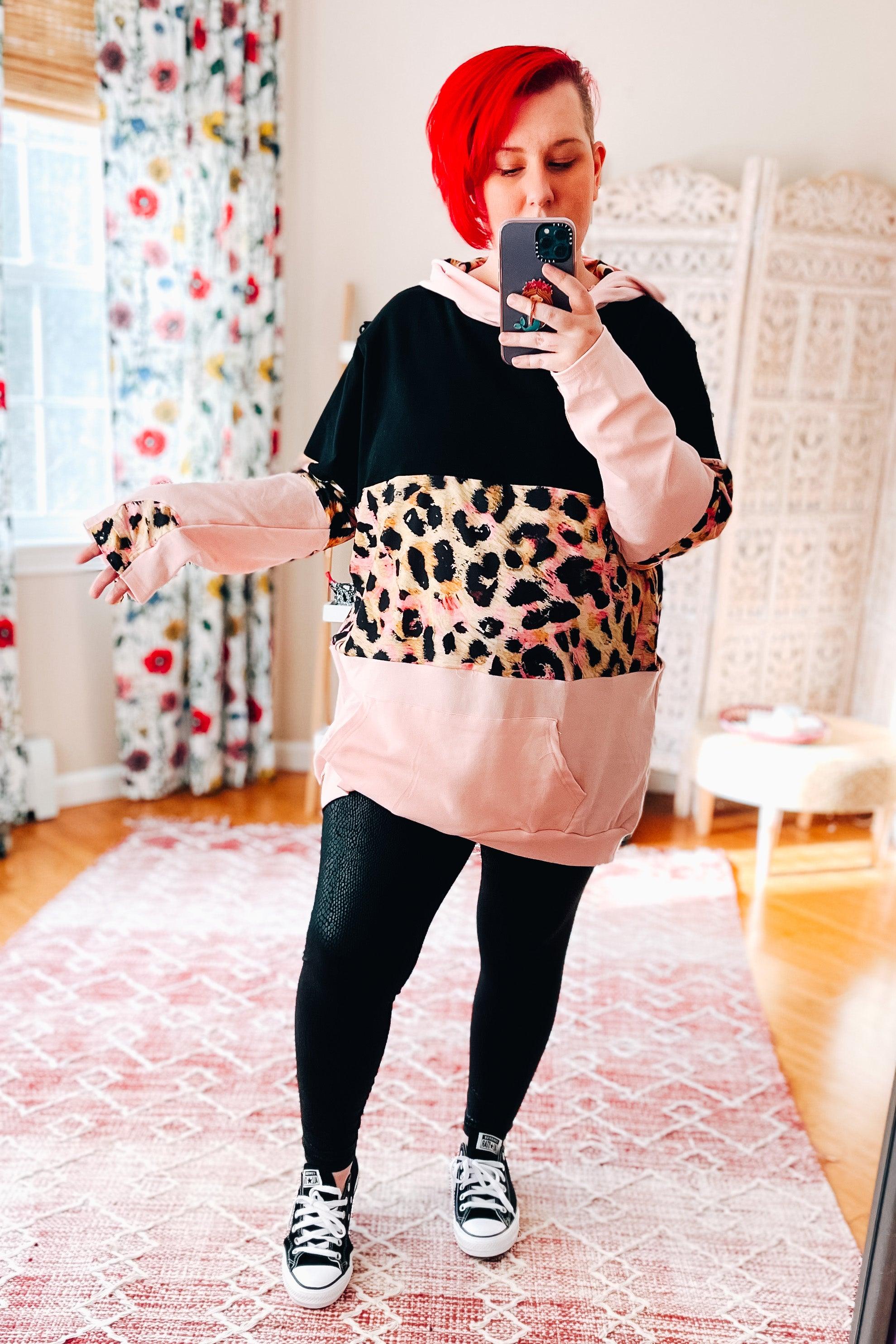 Woman with bright red hair wearing color-blocked hoodie with leopard print and light pink details taking mirror selfie indoors