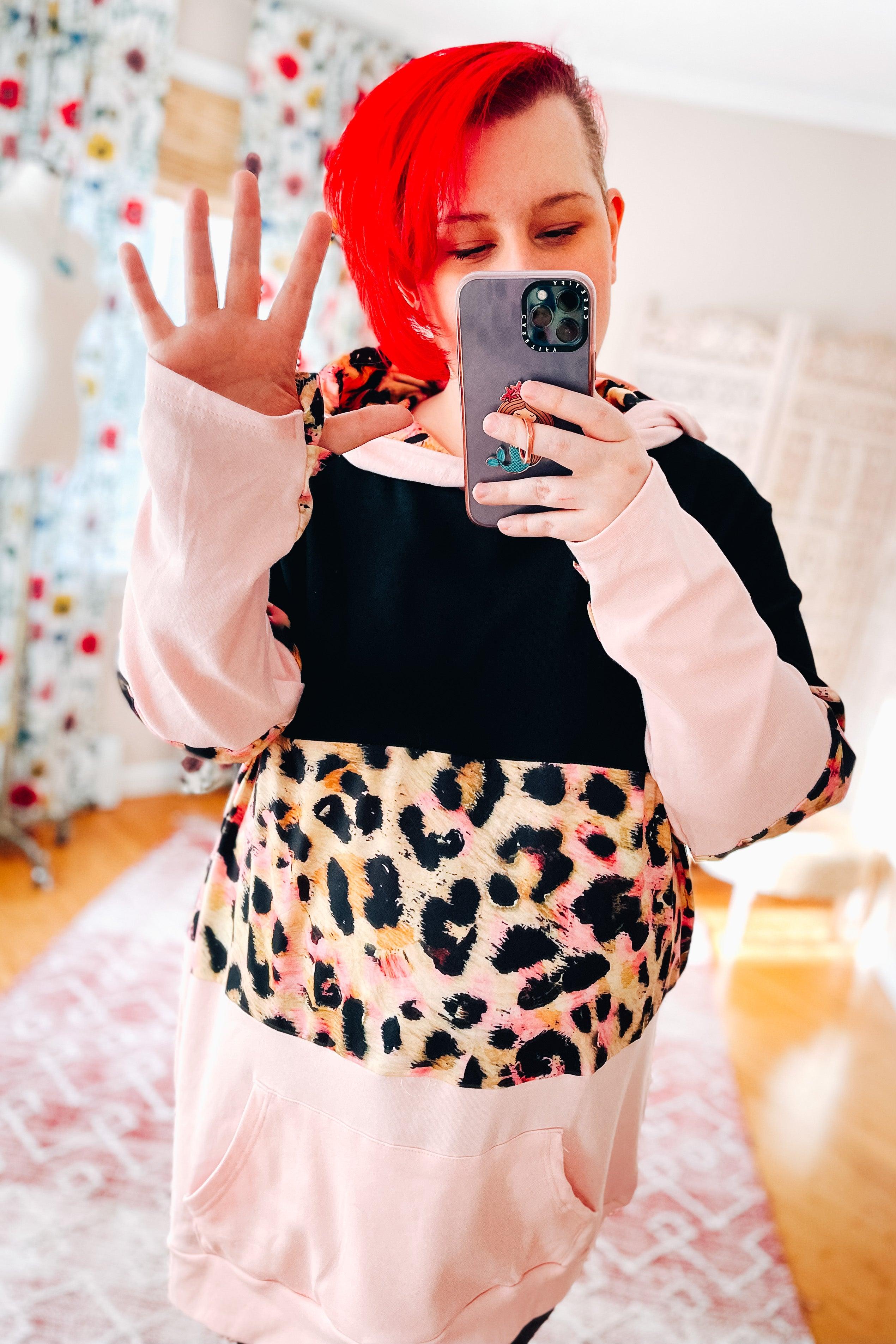 Woman with bright red hair wearing black and pink leopard print Your Song Sweetheart Hoodie indoors taking selfie