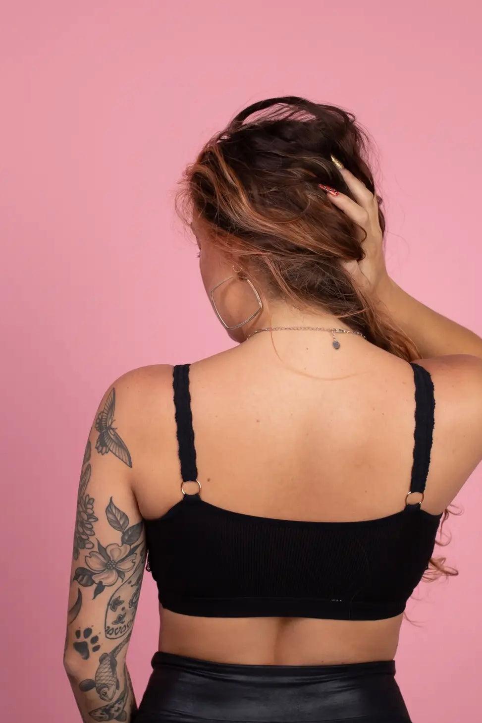 Back view of woman wearing black lace trim bralette with braided straps and floral tattoos on arm against pink backdrop