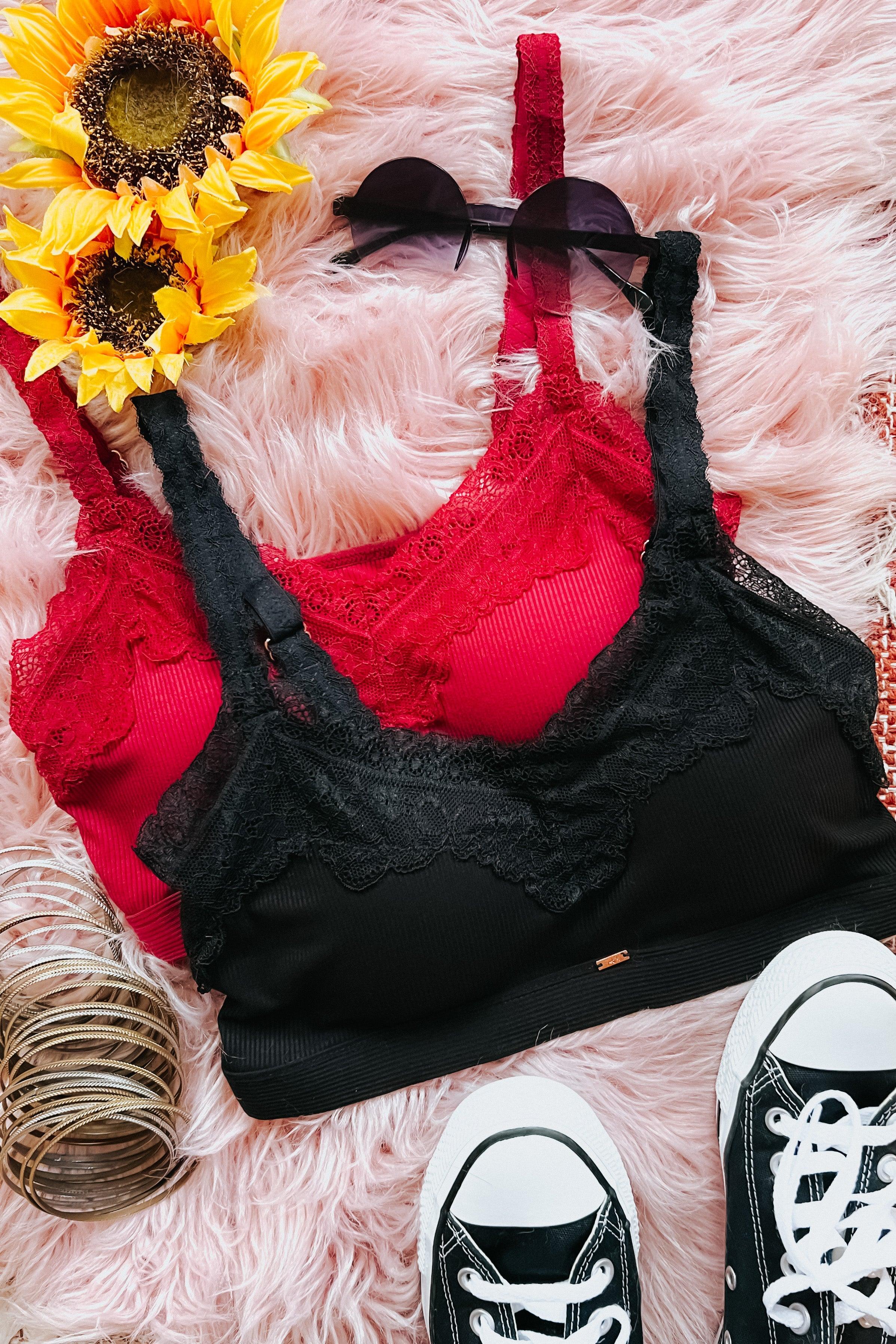 Two lace-trimmed bralettes in black and red on fluffy pink surface with sunglasses, bracelets, sunflowers, and black sneakers
