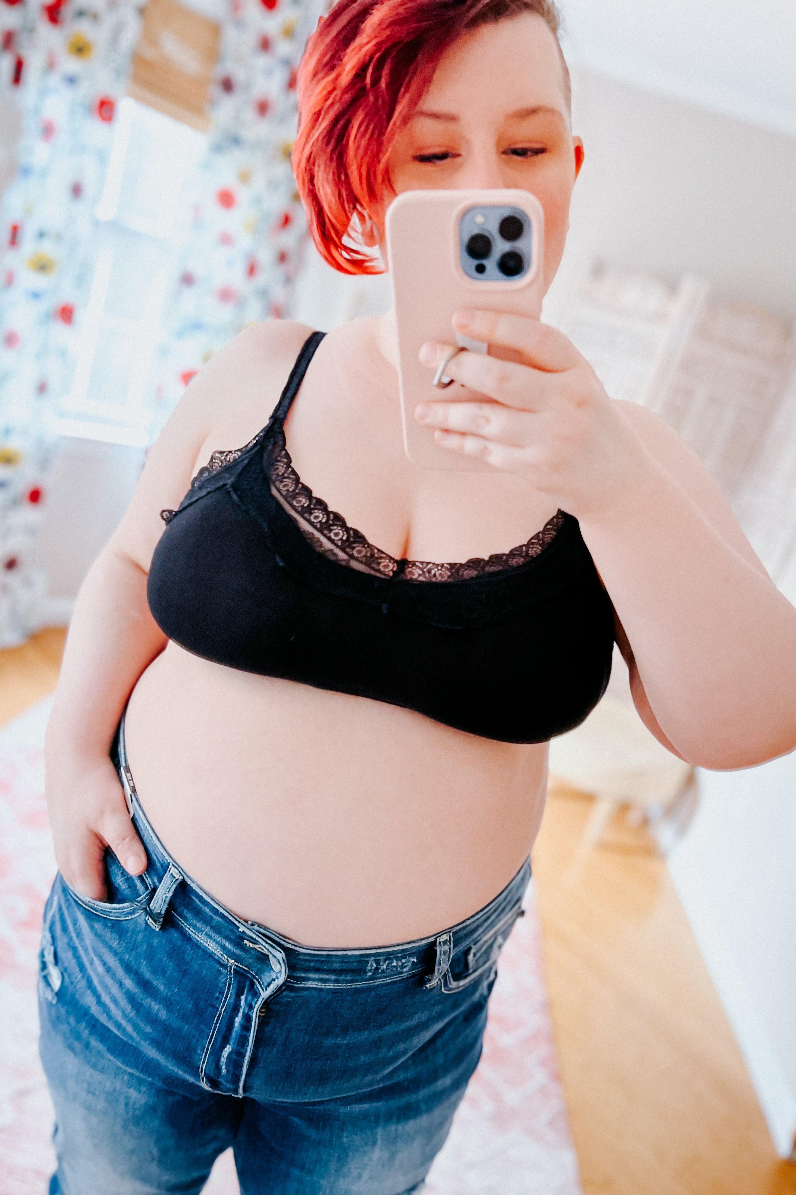 Plus-size woman wearing black lace-trim bralette and unbuttoned jeans taking mirror selfie in bright room