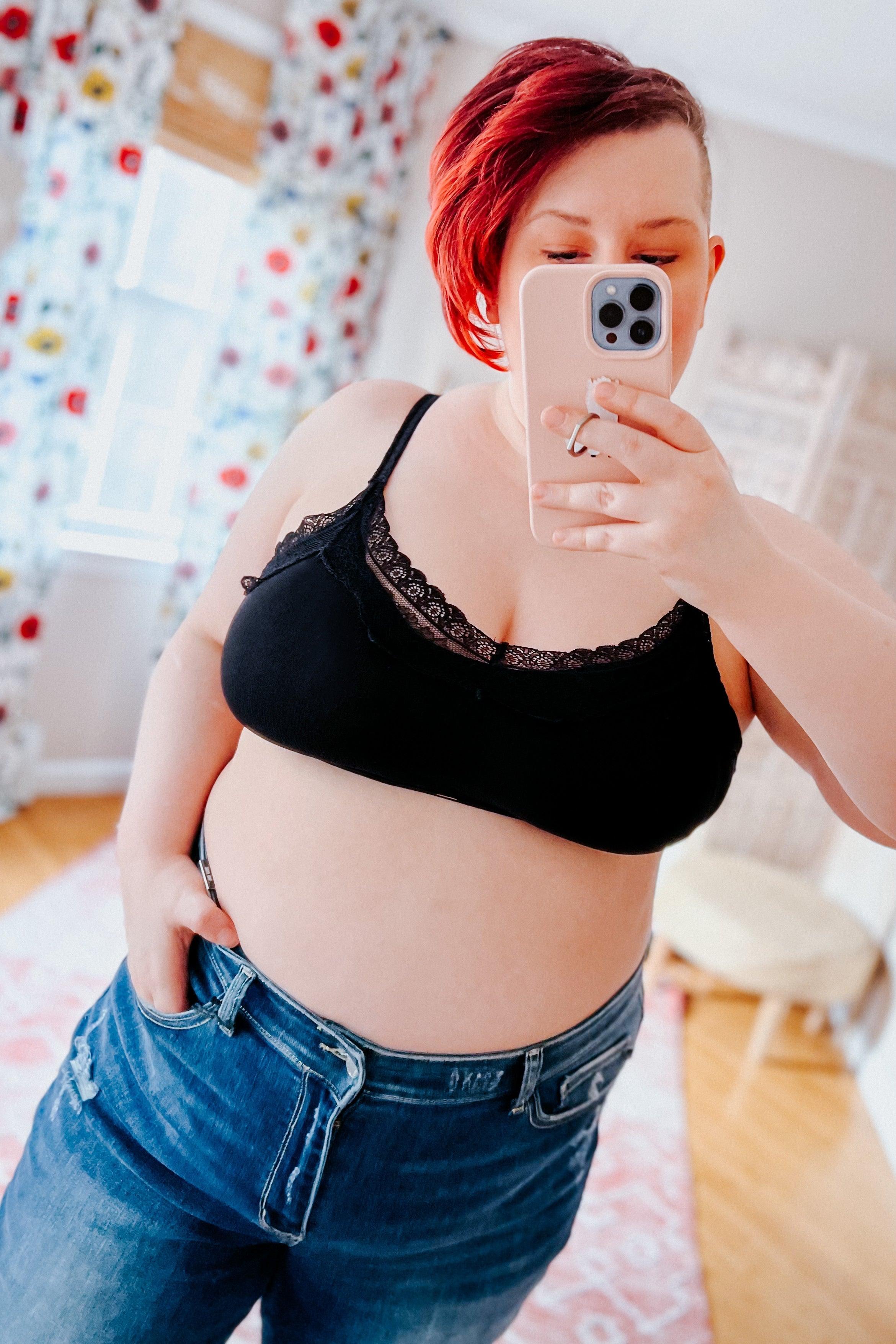 Woman with red hair wearing black lace-trim bralette and unbuttoned jeans taking a mirror selfie indoors with floral curtains