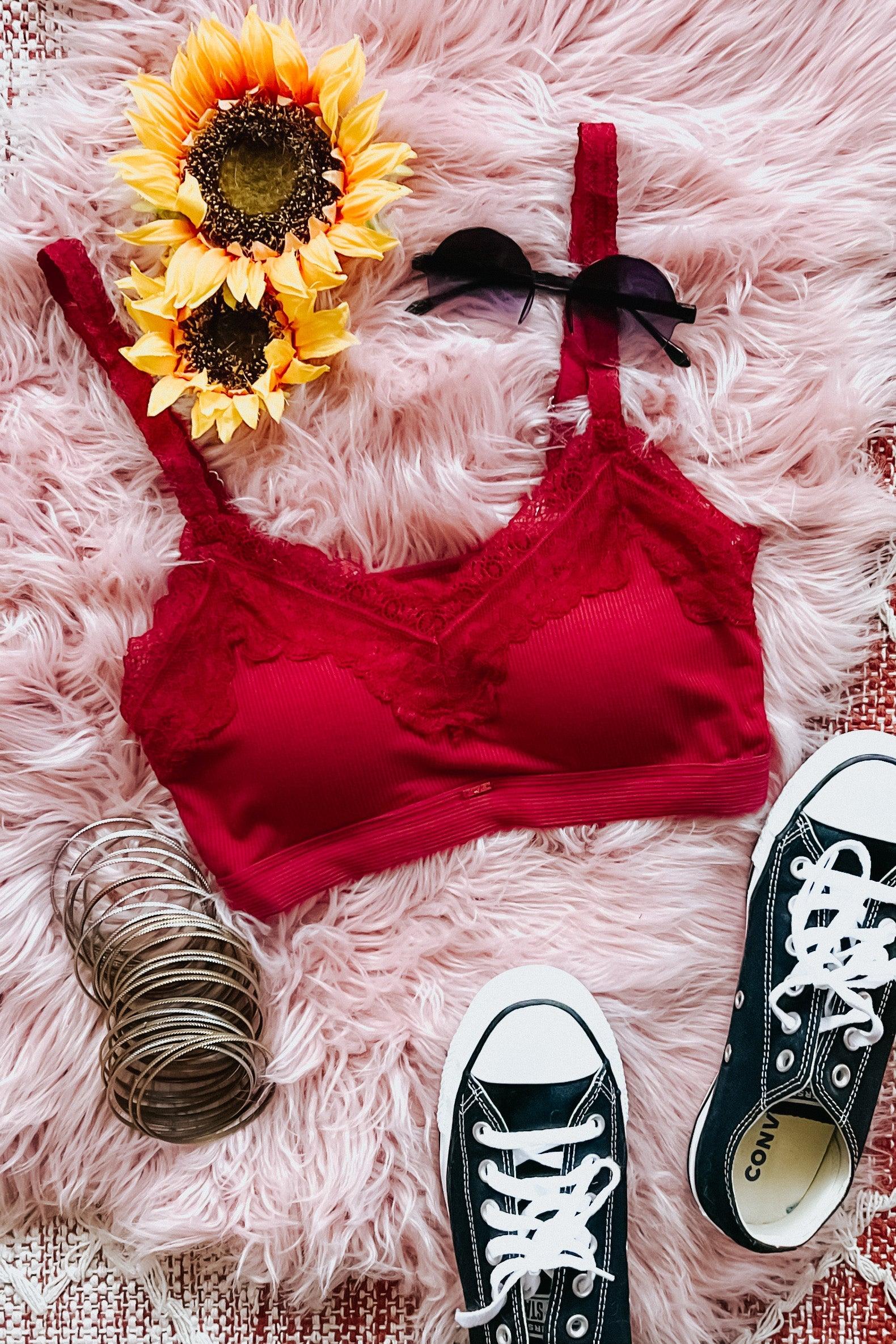 Wine red lace-trim bralette styled with black sunglasses, sunflower bouquet, silver bangles, and black sneakers on pink fur background
