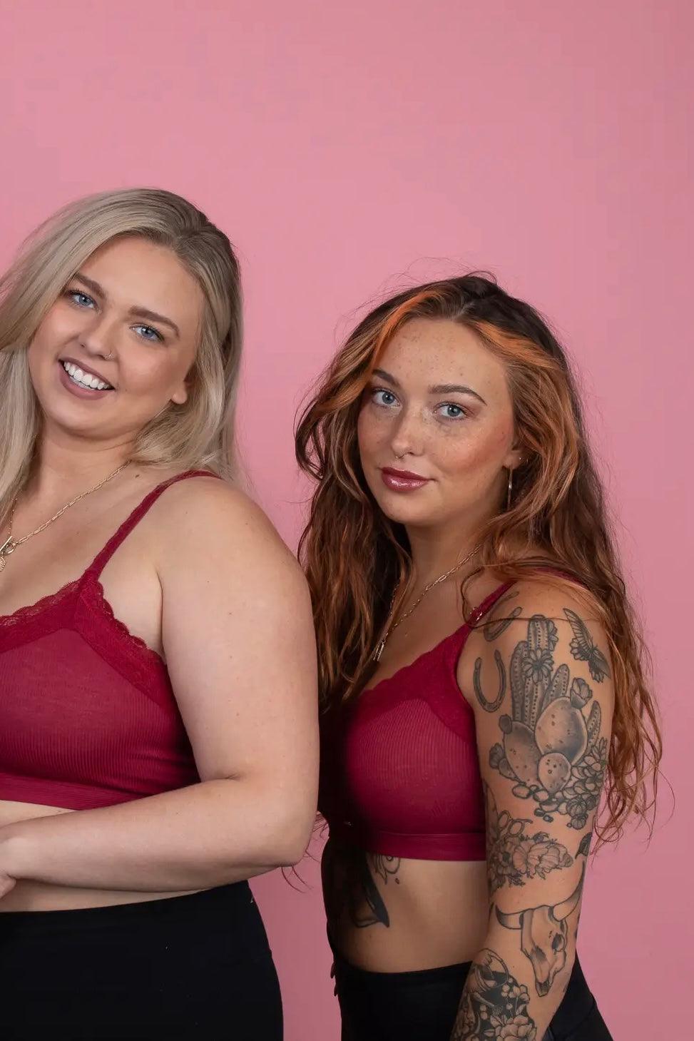 Two women wearing wine-colored lace-trim bralettes posing against a pink backdrop with bold tattoos and natural makeup