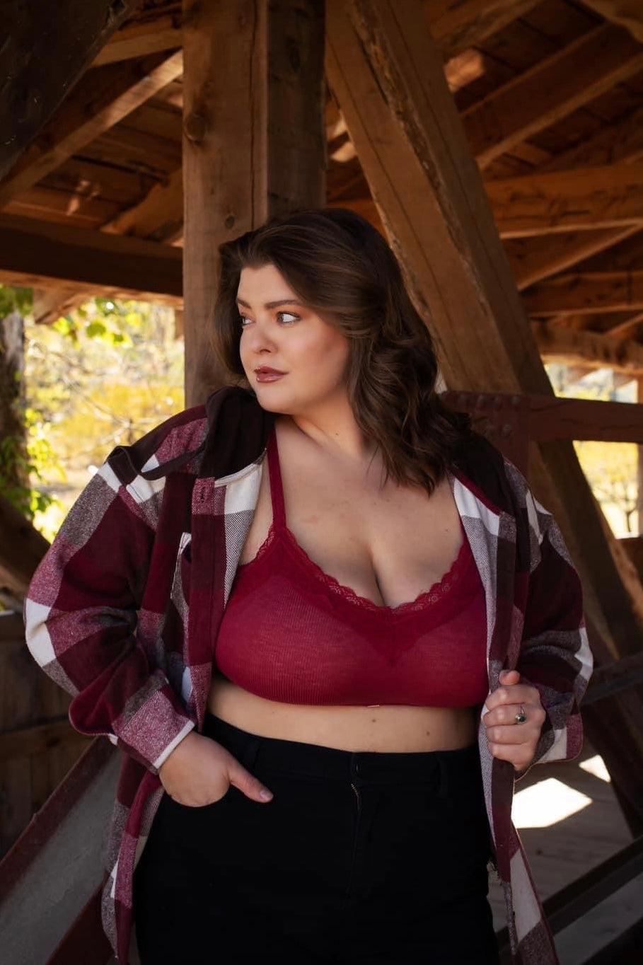 Plus-size woman wearing wine-colored lace trim bralette under plaid jacket, standing in rustic wooden structure outdoors
