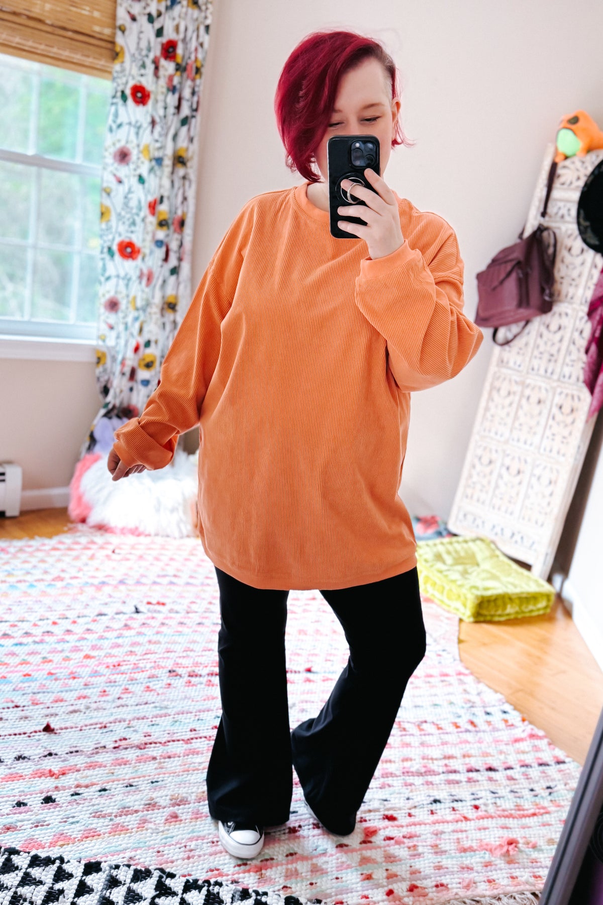 Red haired woman modeling an oversized orange corded sweatshirt with black flared pants and sneakers in a bright bedroom mirror selfie