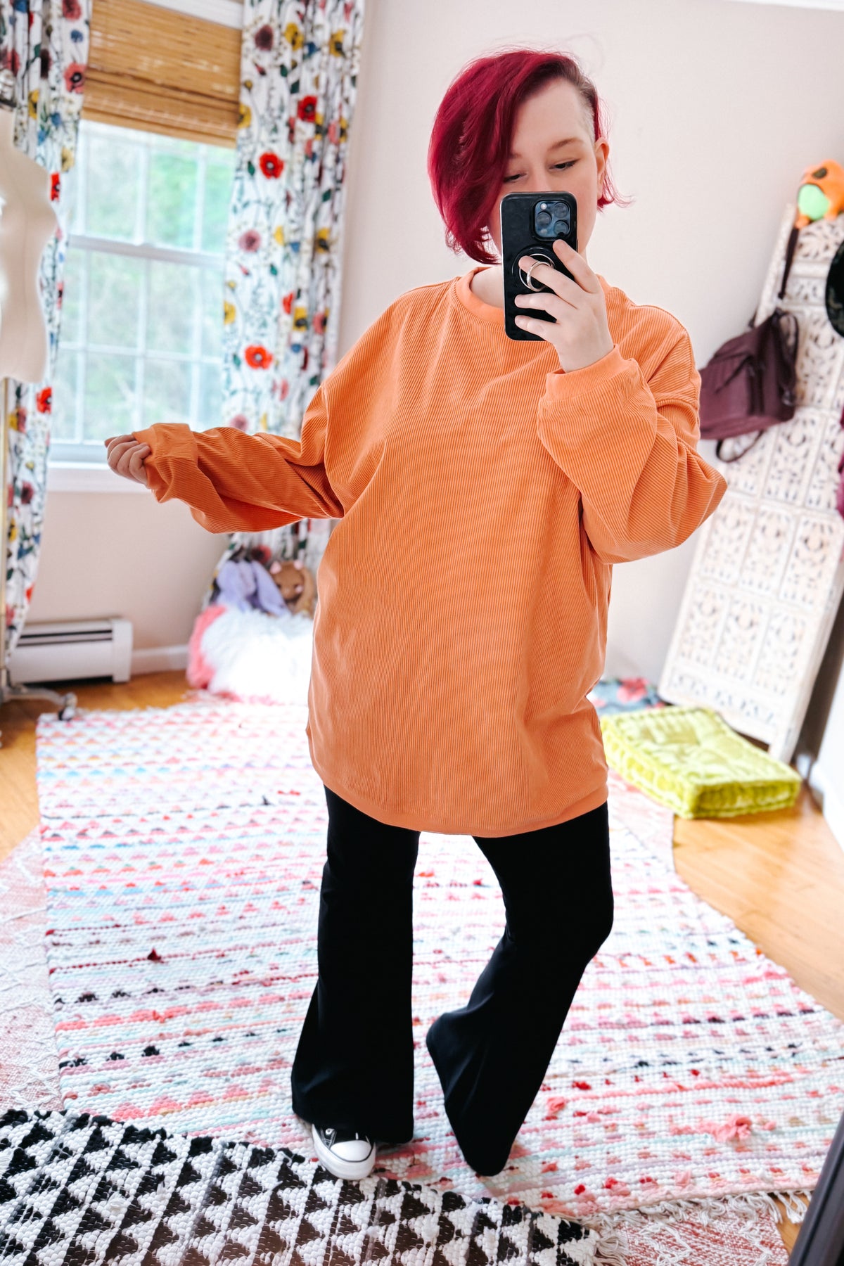 Red haired woman taking a mirror selfie in an oversized orange corded sweatshirt with black pants and sneakers in a colorful bedroom