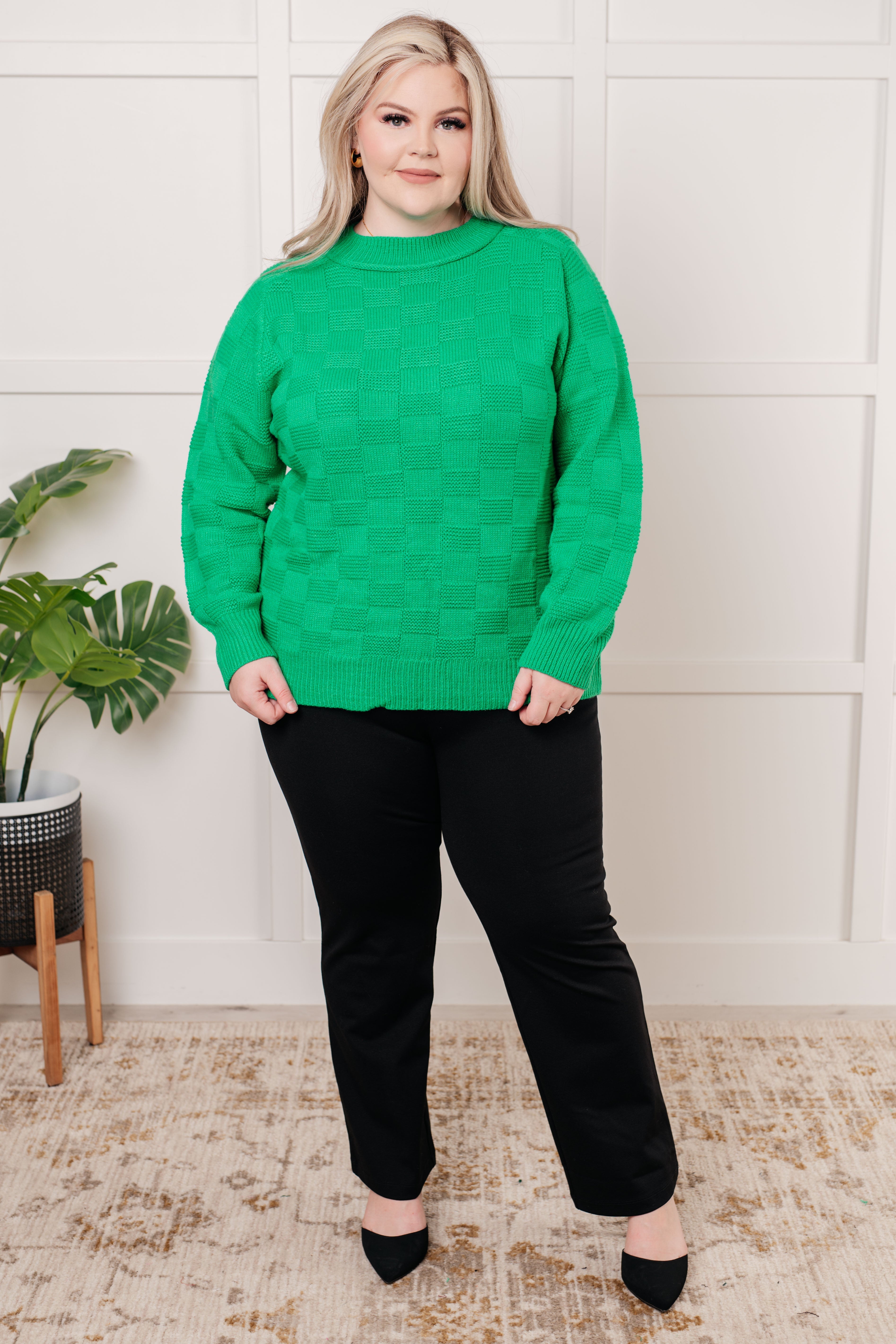 Woman wearing a green sweater, black pants and black heels standing in a room with white wall, a plant and rug.