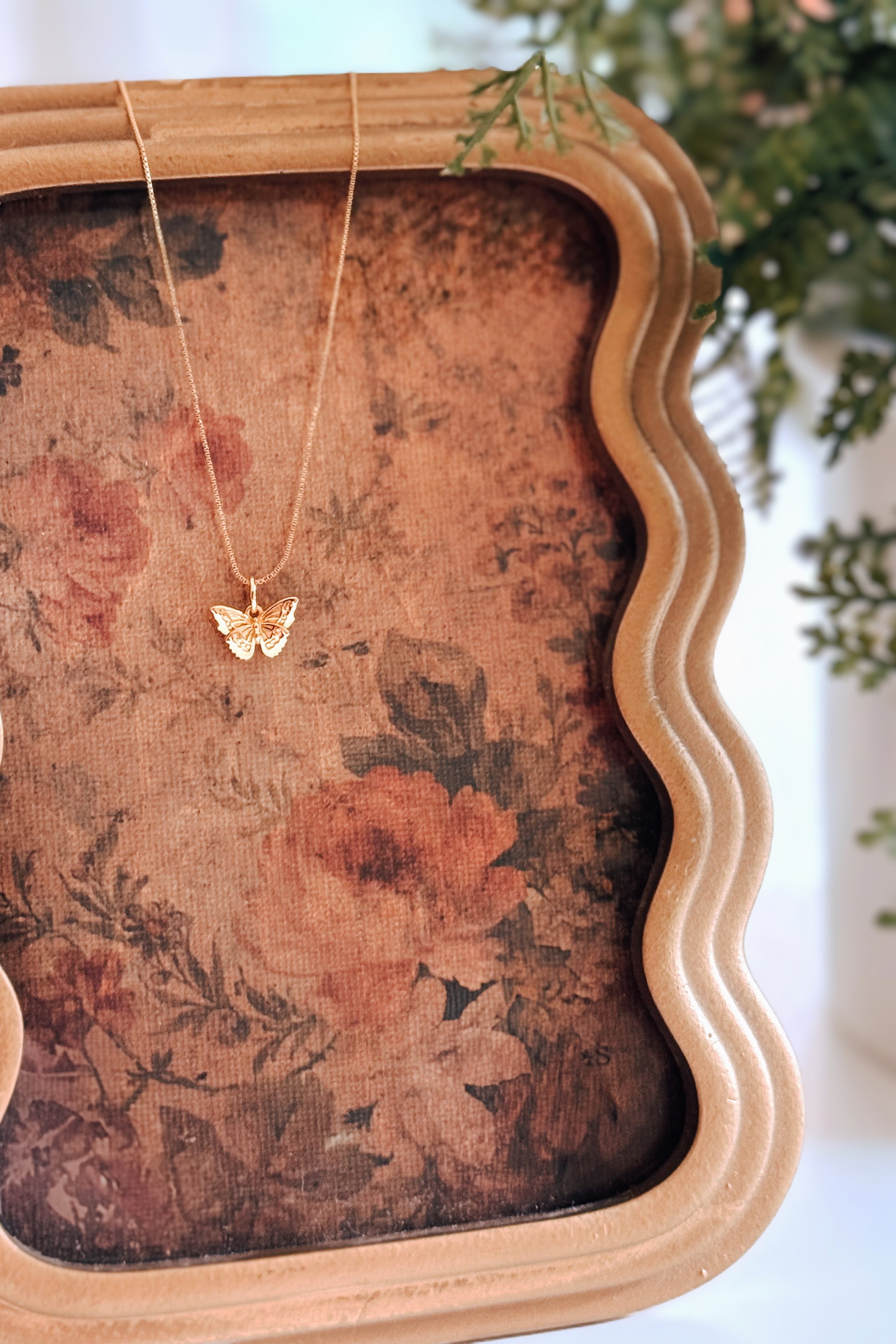 Gold butterfly pendant necklace hanging on floral fabric inside a scalloped wooden frame in soft light