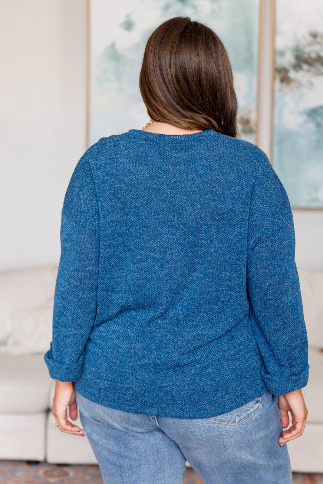 Woman with her back facing the camera wearing a blue sweater and jeans in a room with a painting on the wall.