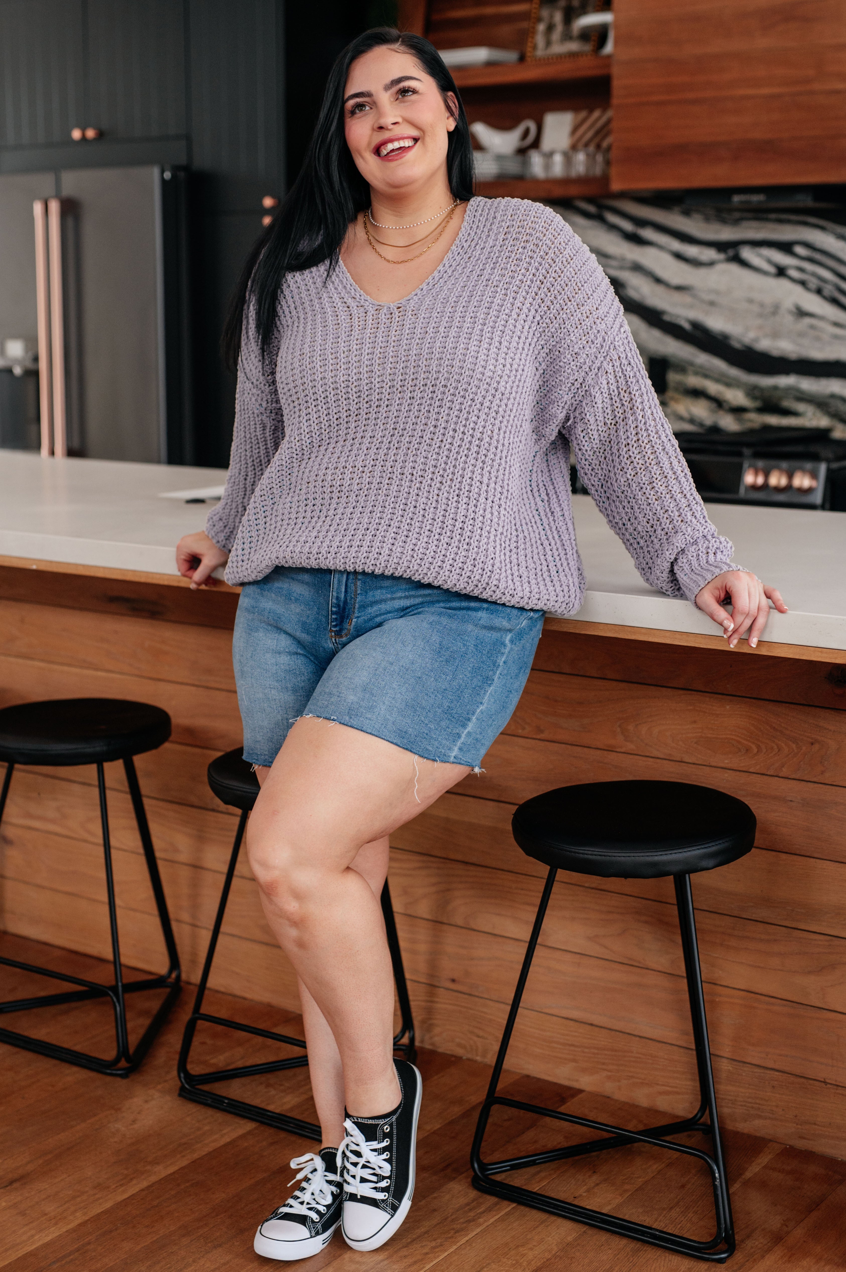 Woman in a kitchen wearing a purple sweater and denim shorts, standing on a stool.