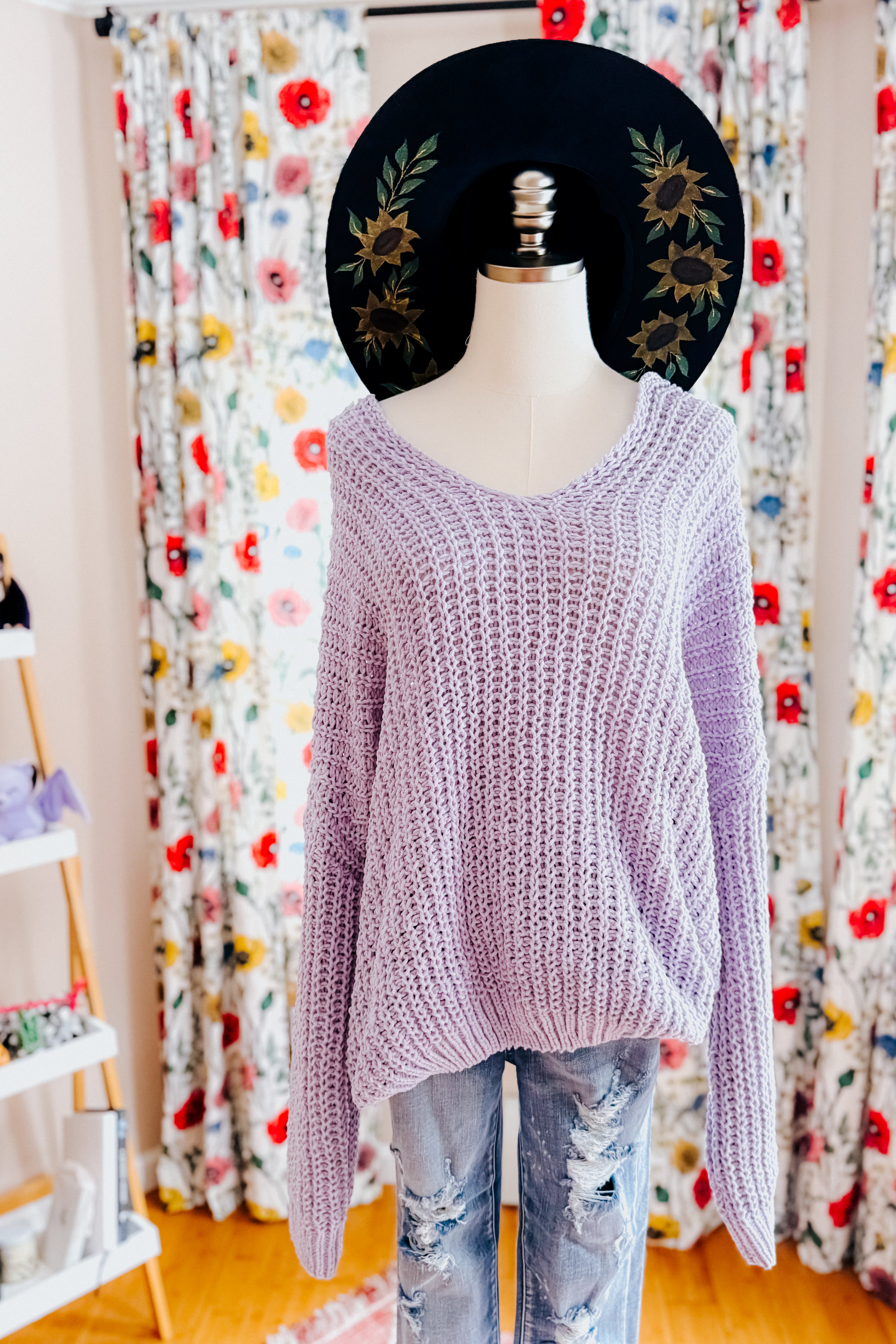A dress form wearing a chunky lavender sweater, blue jeans and a wide brim painted had in an indoor setting.