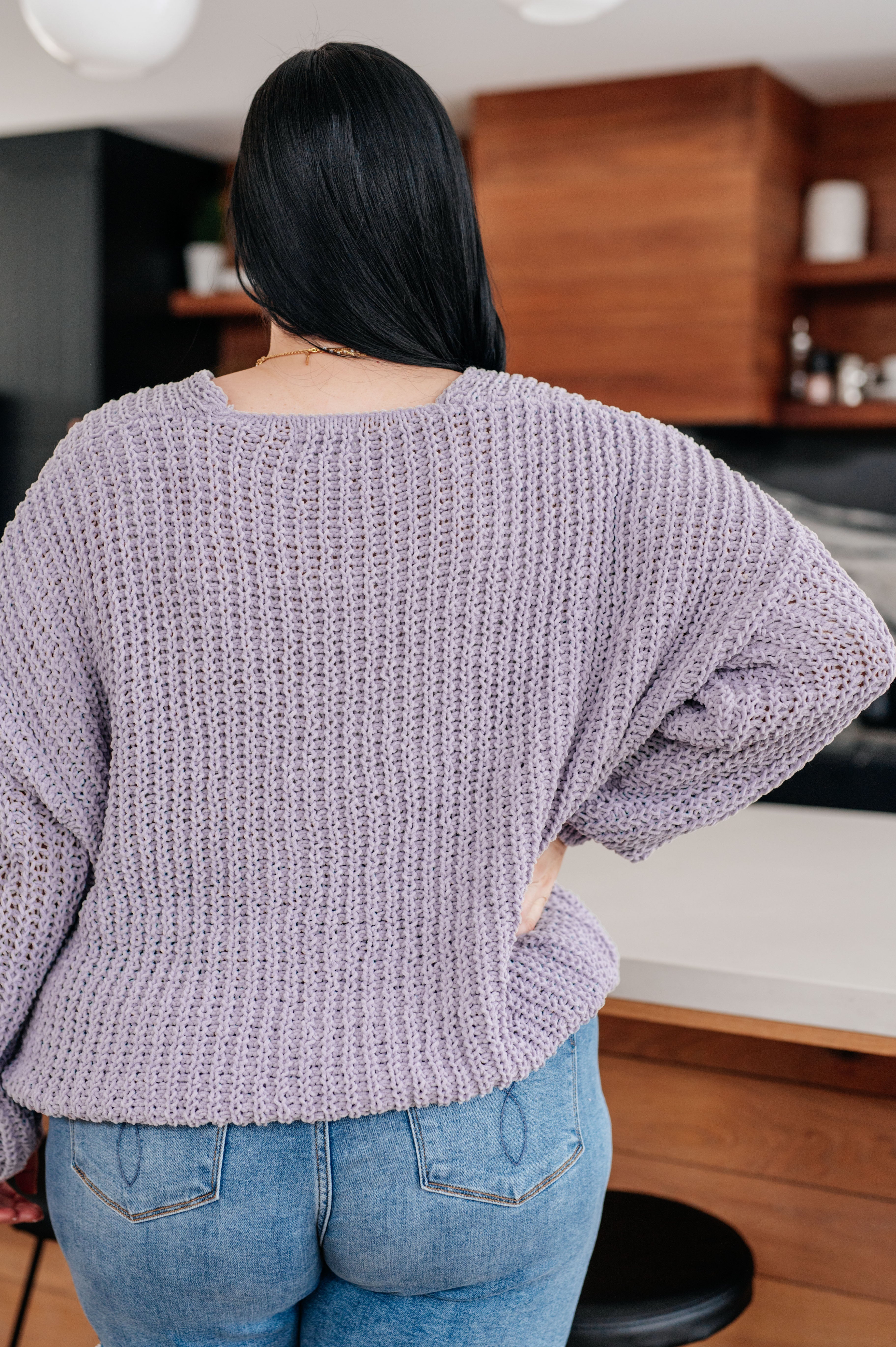 Woman with her back facing the camera wearing a purple sweater and blue jeans in a kitchen setting