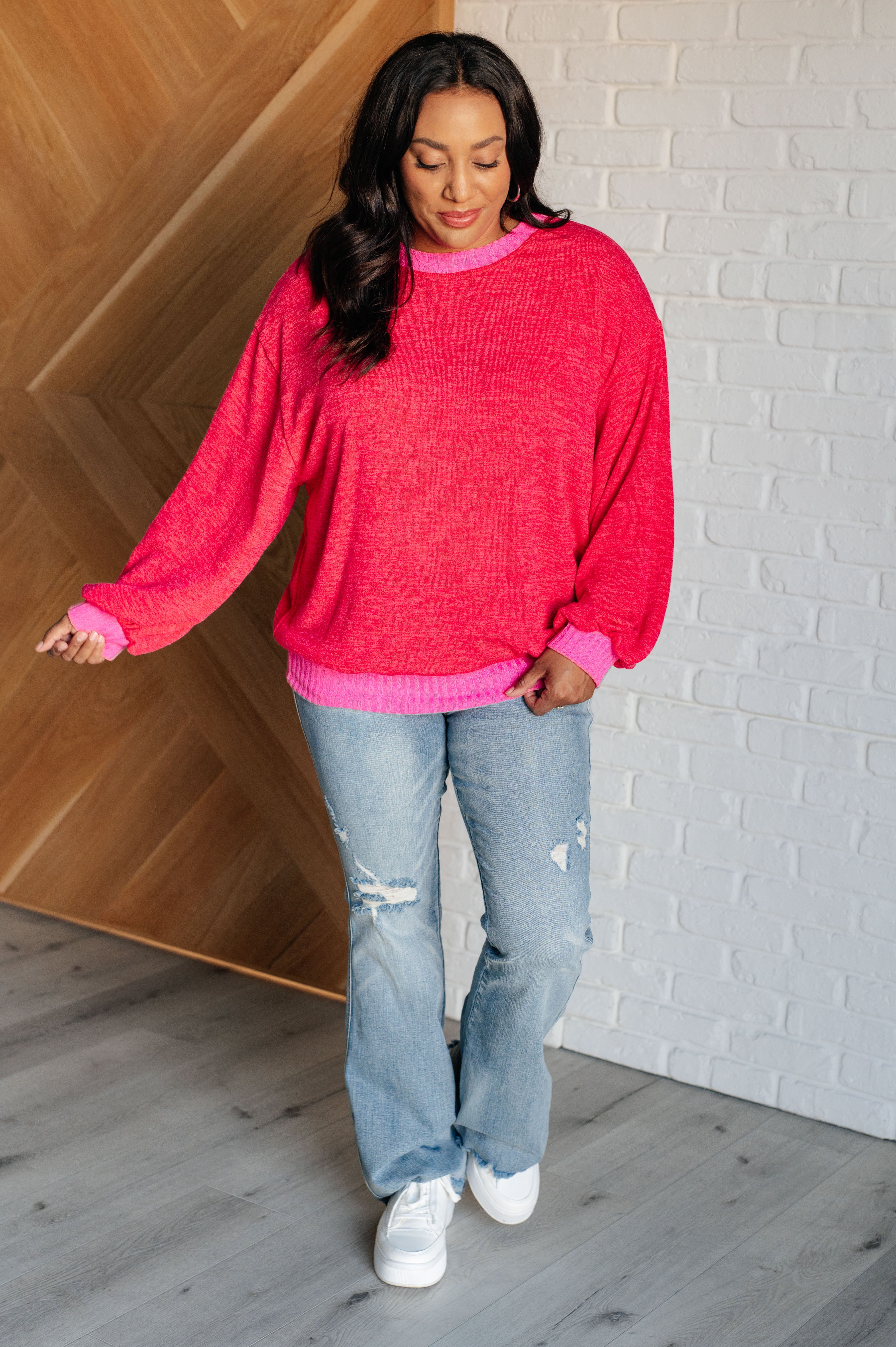 Woman wearing a red sweater with bright pink trim and blue jeans standing against a white brick wall background