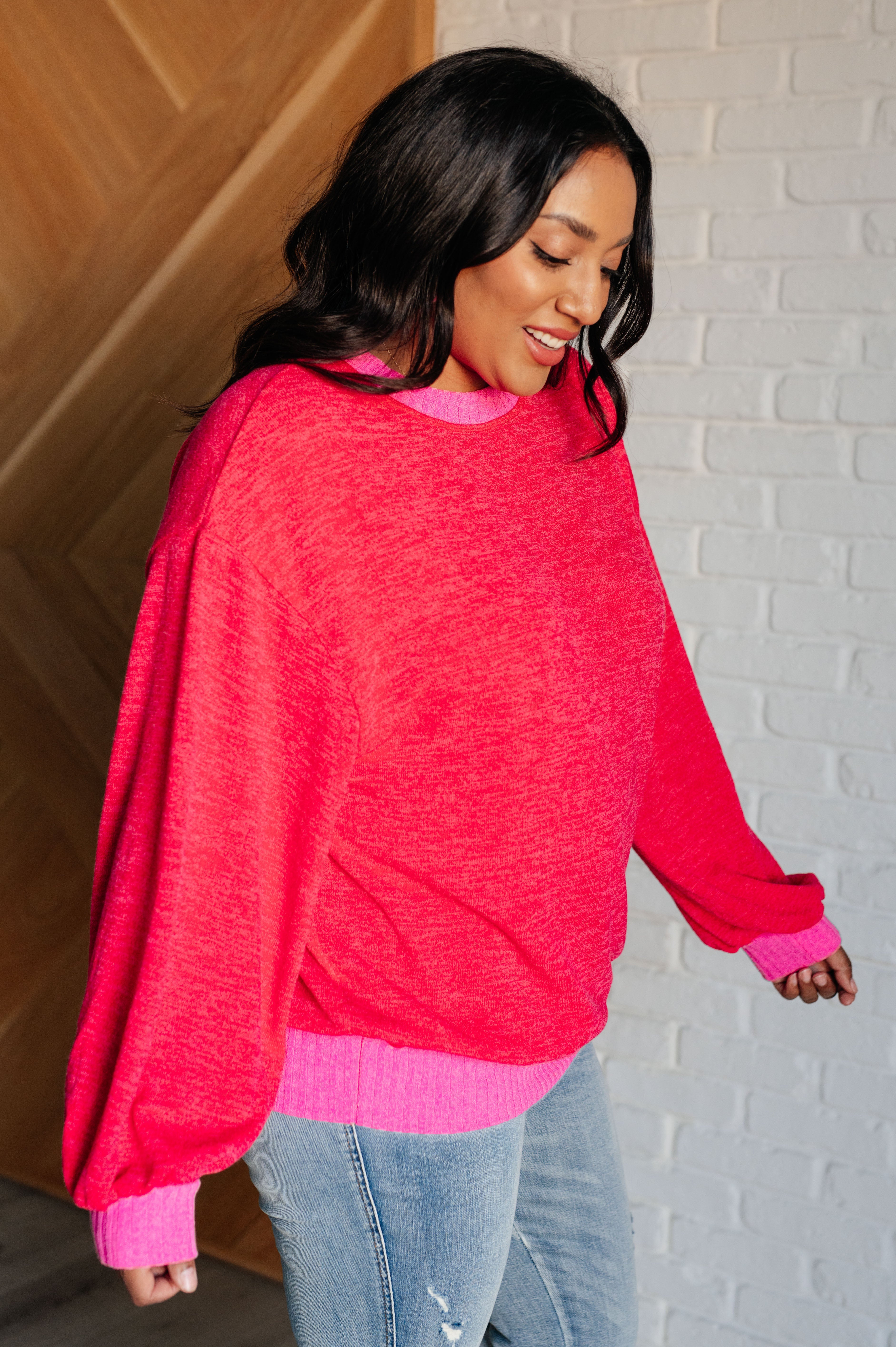 Woman wearing a red sweater with bright pink trim and blue jeans standing against a white brick wall background