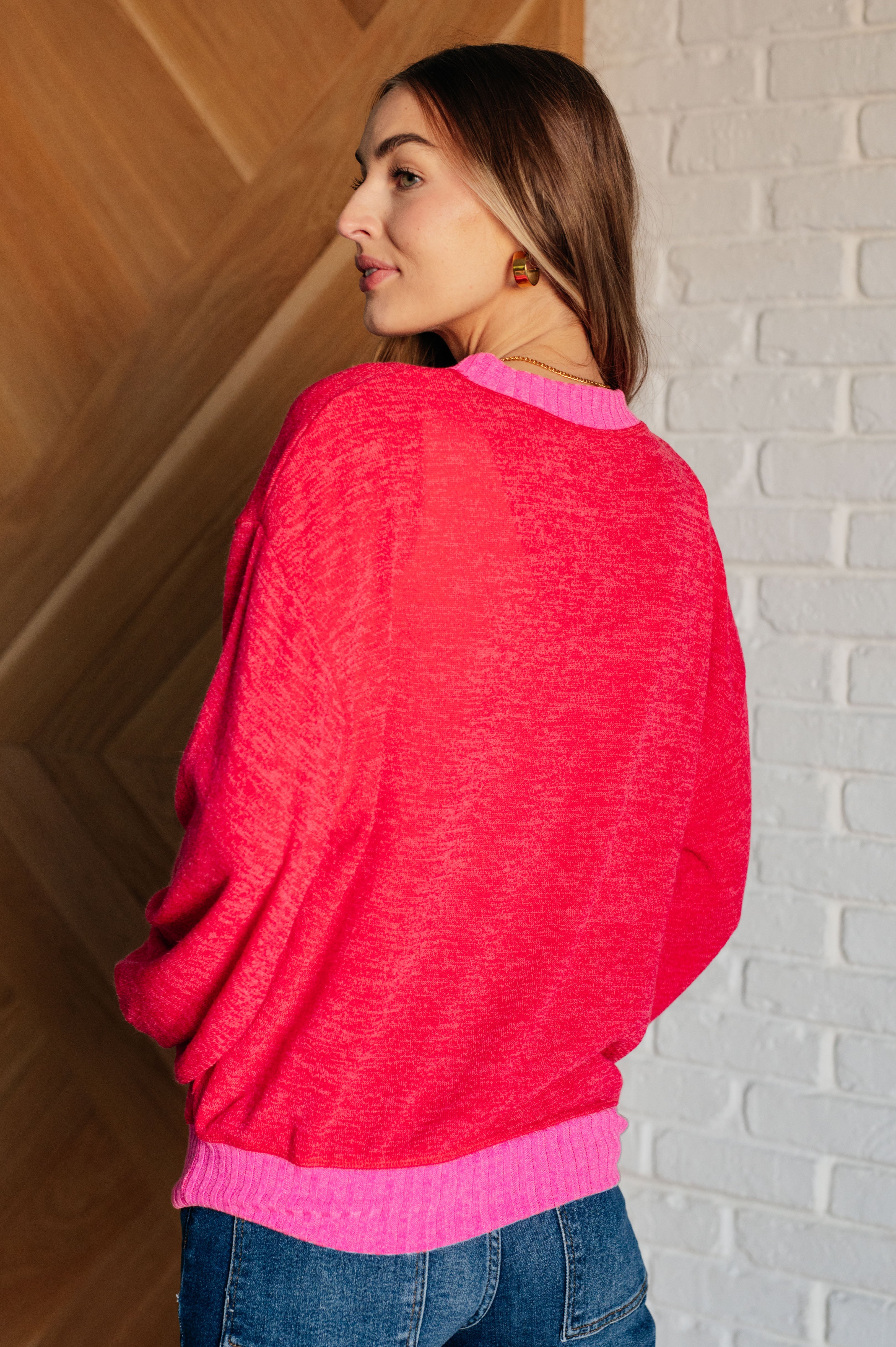 Woman wearing a red sweater with bright pink trim and blue jeans standing against a white brick wall background