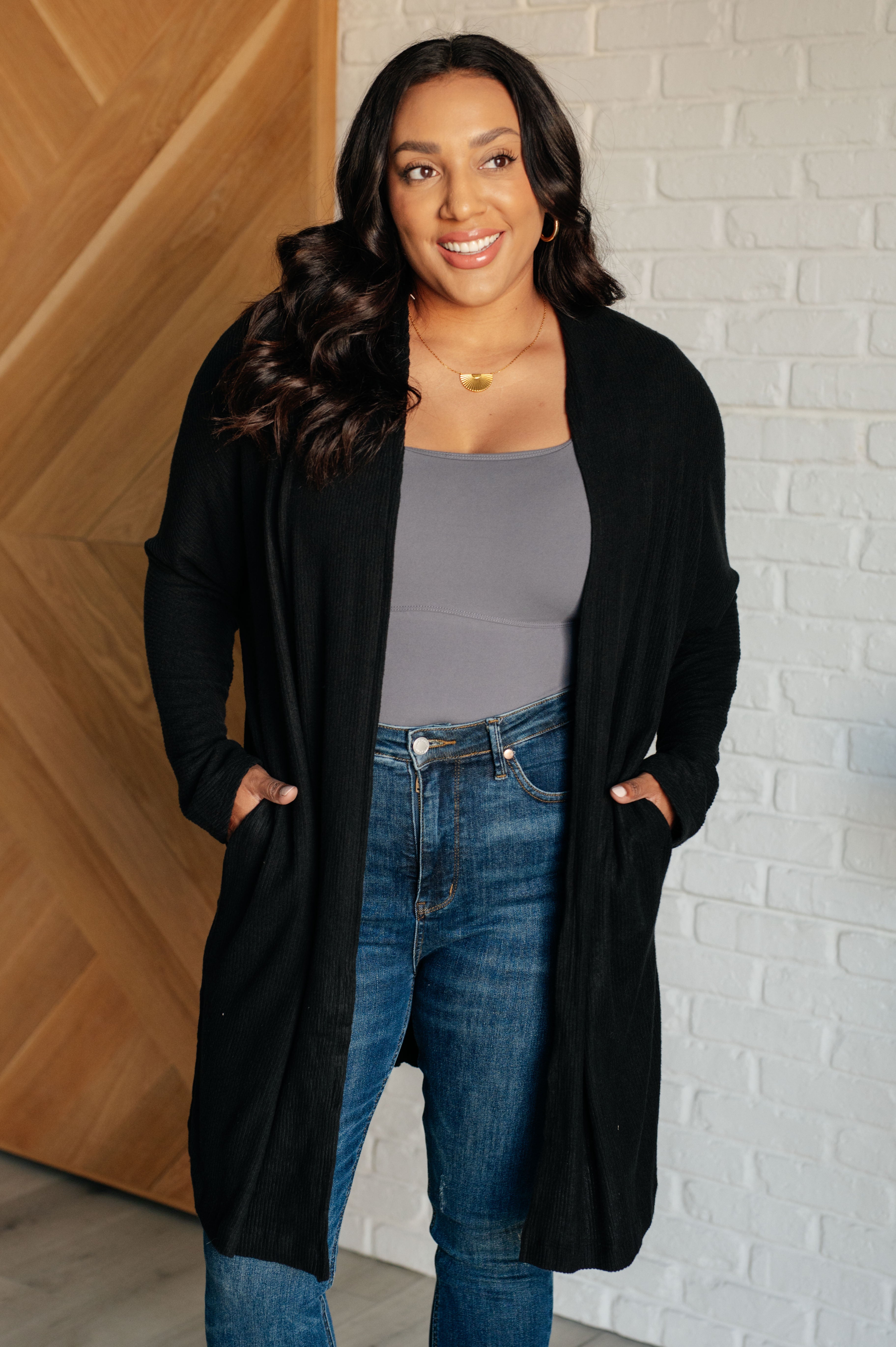 Woman wearing a black cardigan, gray top, and blue jeans standing against a brick wall.