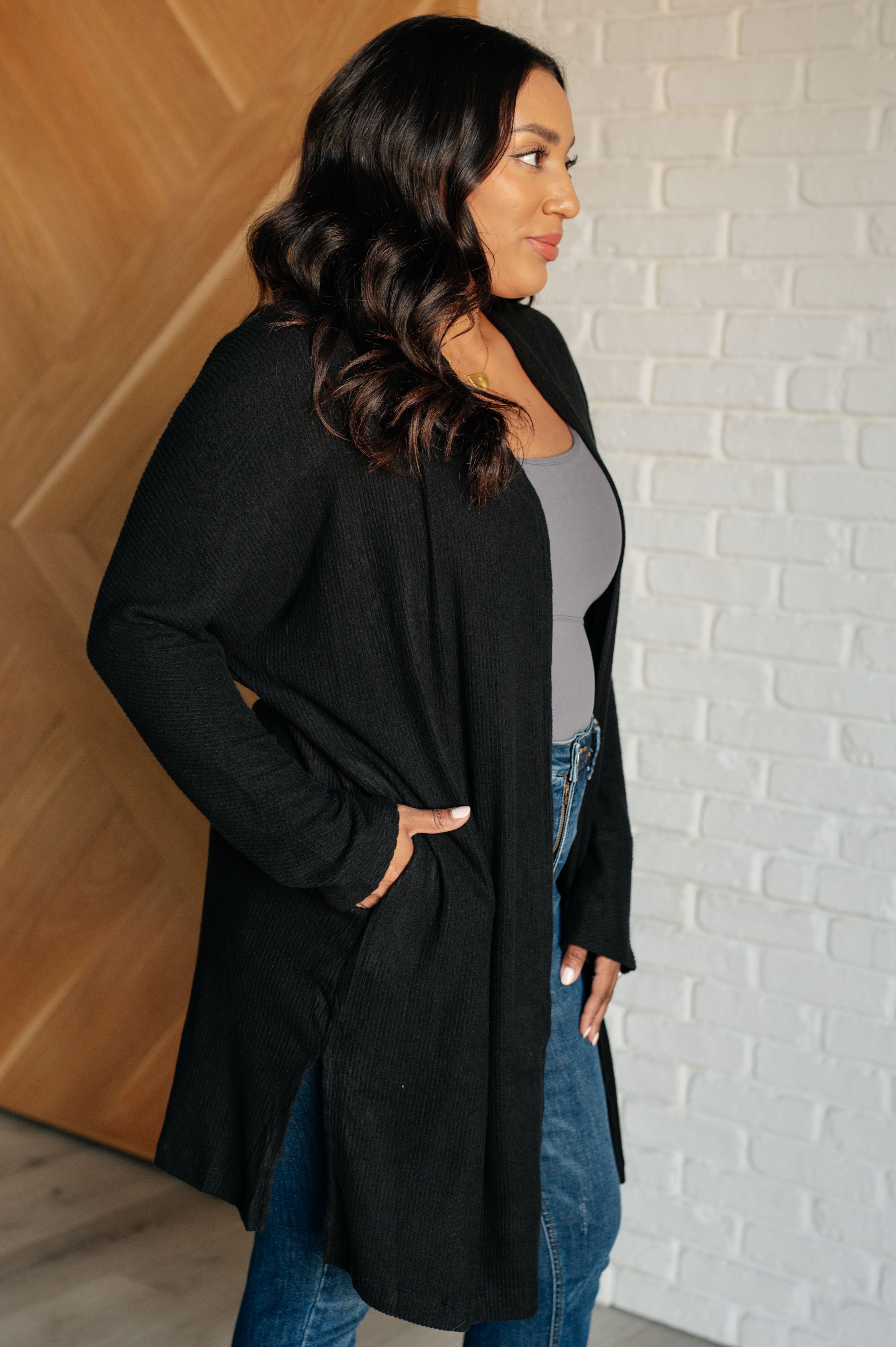 Woman wearing a black long cardigan, a grey top and blue jeans against a white brick wall.