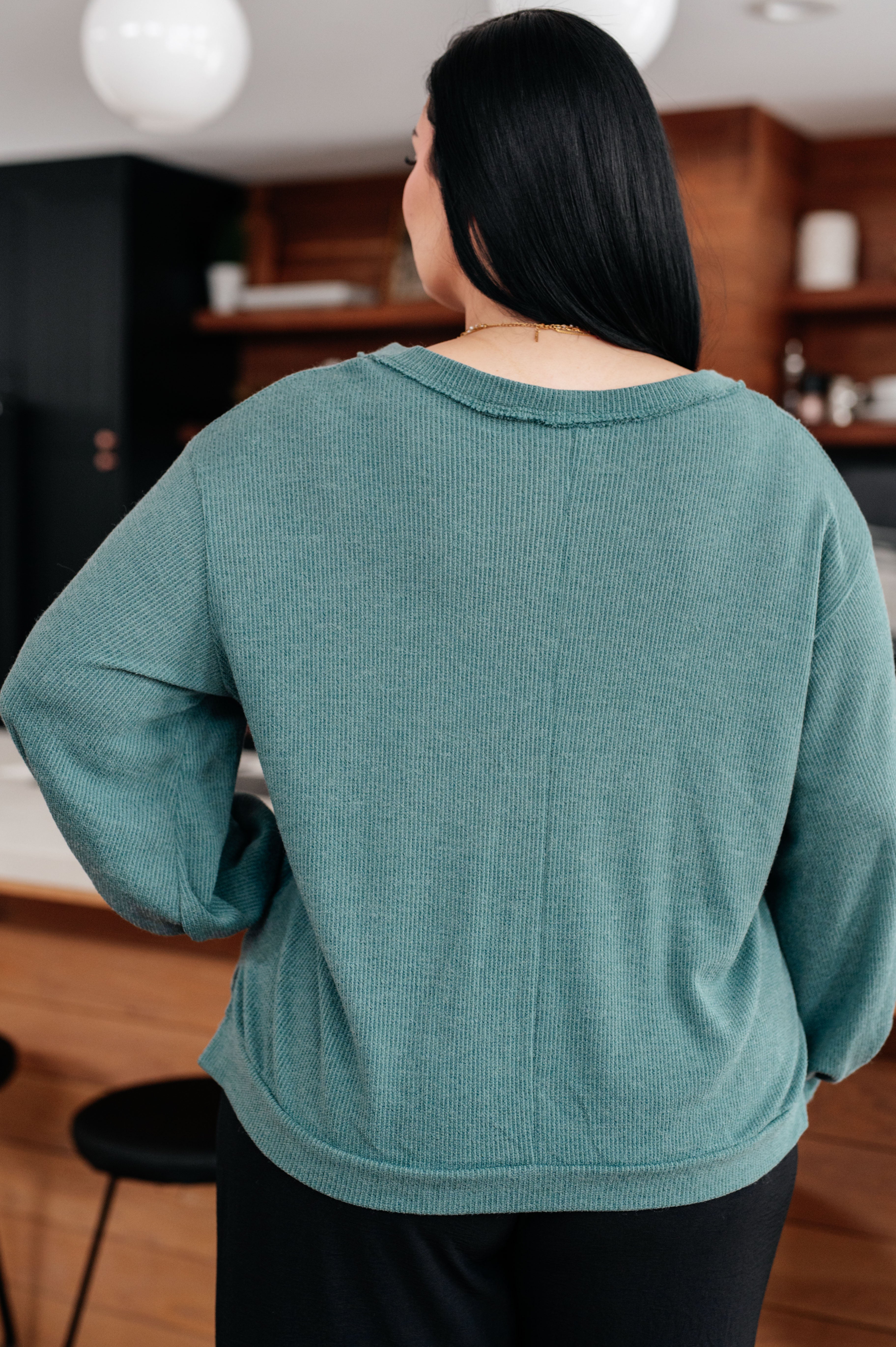 Woman with her back facing the camera wearing a teal sweater and black pants in an indoor setting