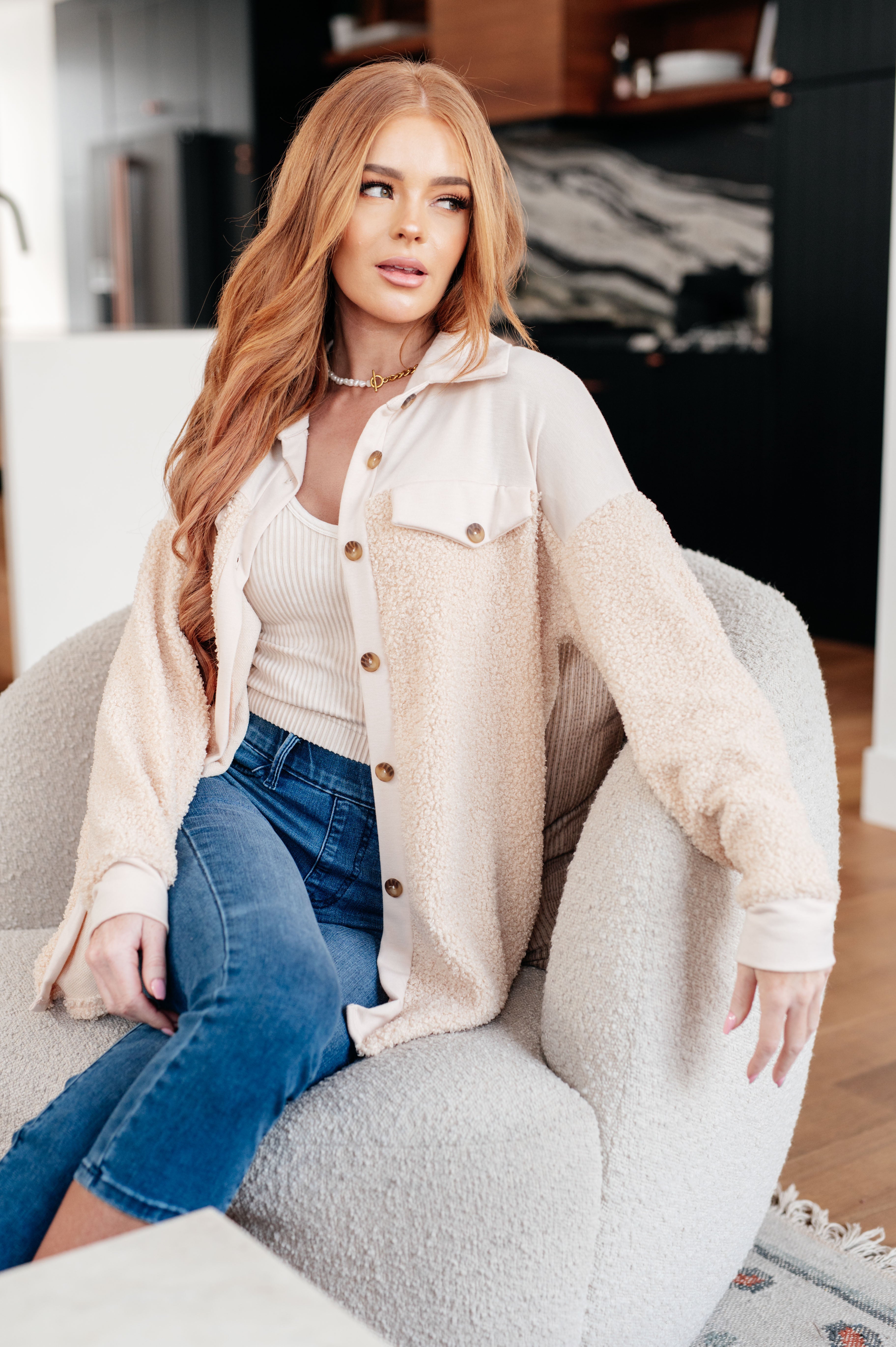 Woman wearing a beige shacket, a beige tank and blue jeans in a cozy living room setting