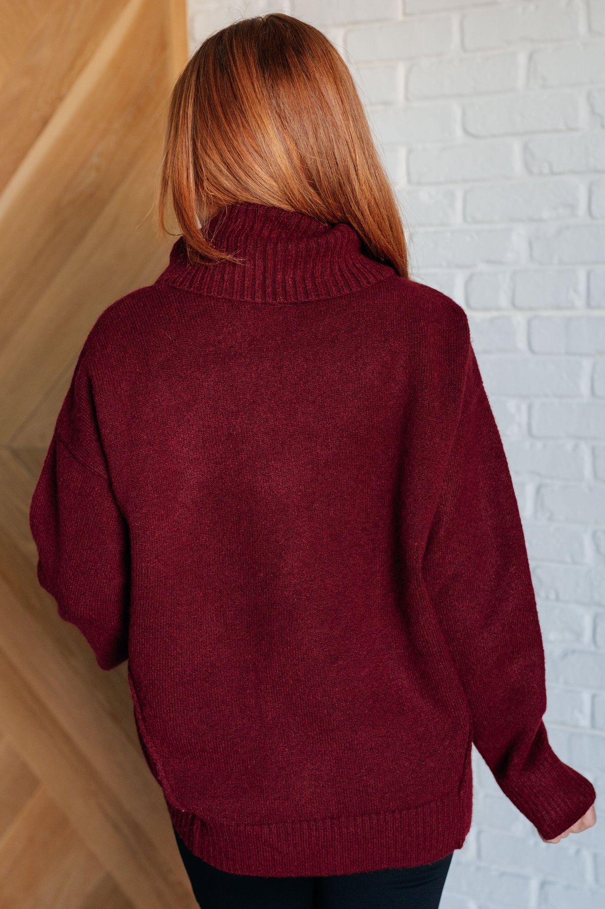 Woman viewed from behind wearing a burgundy turtleneck sweater with a ribbed collar cuffs and hem against a white brick wall