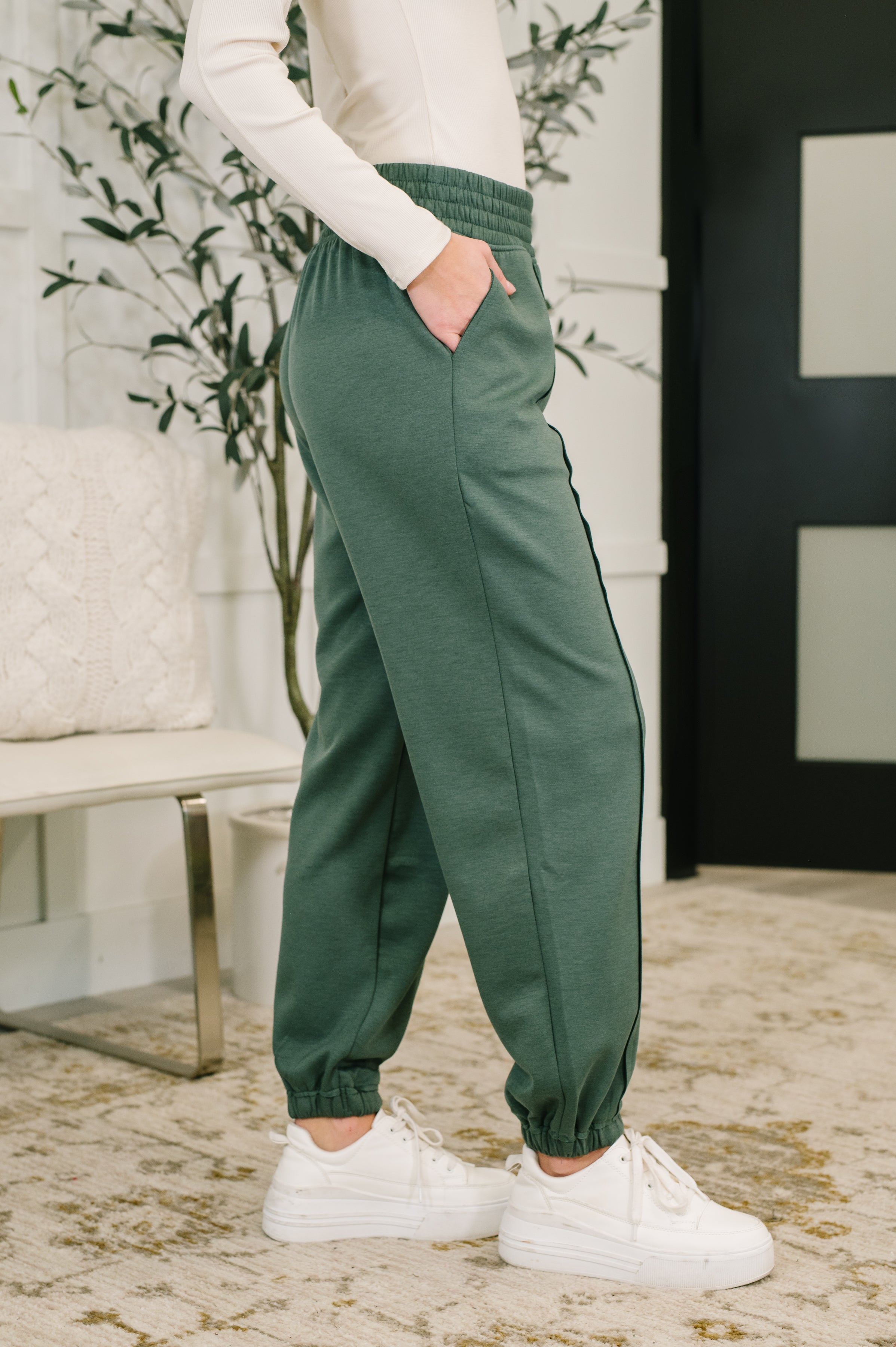 Woman wearing green jogger pants and white sneakers in a room with a plant and bench.