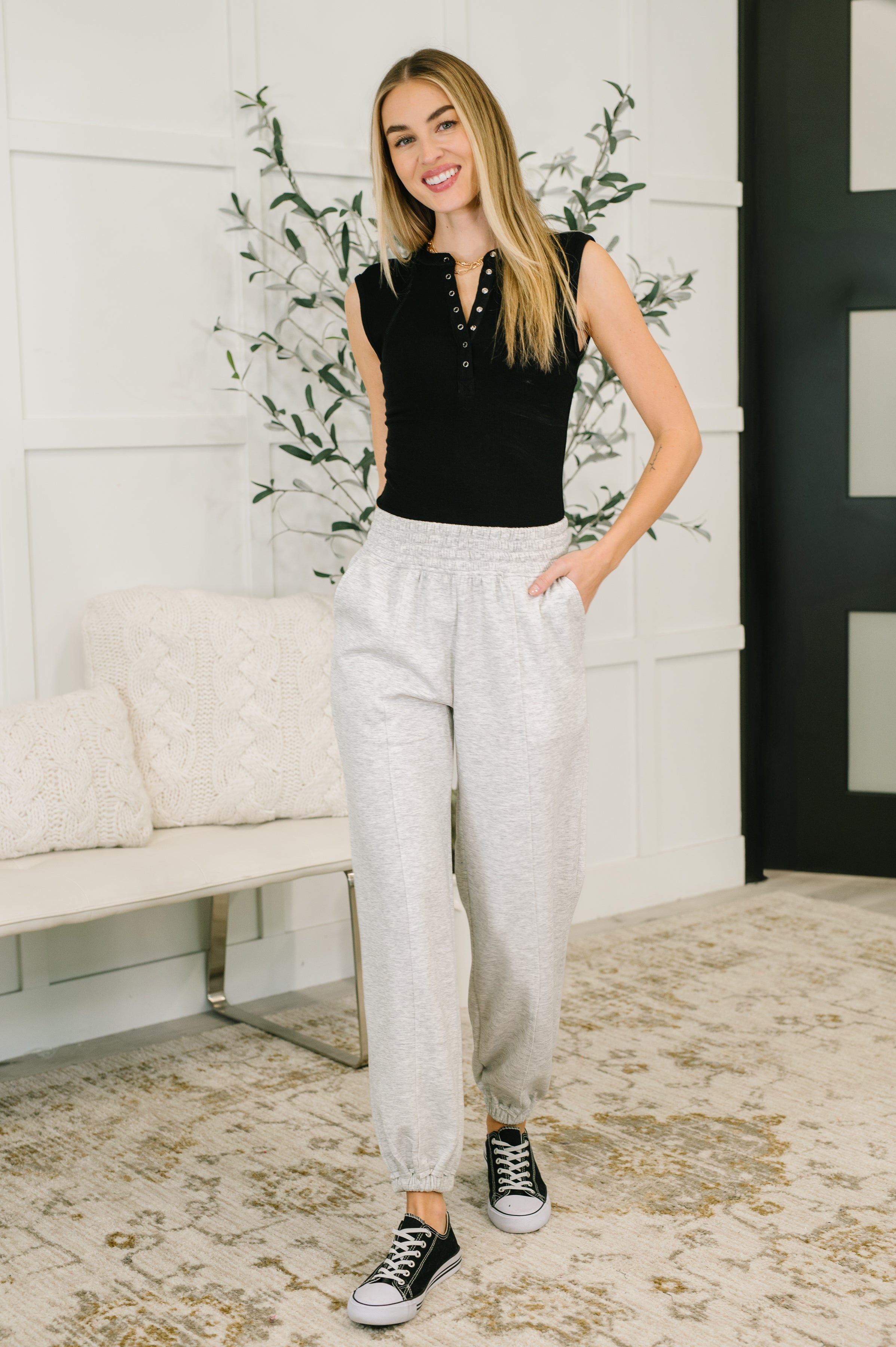 Woman wearing a black top, light gray jogger pants and converse sneakers standing in a room with a white wall and decorative plant.