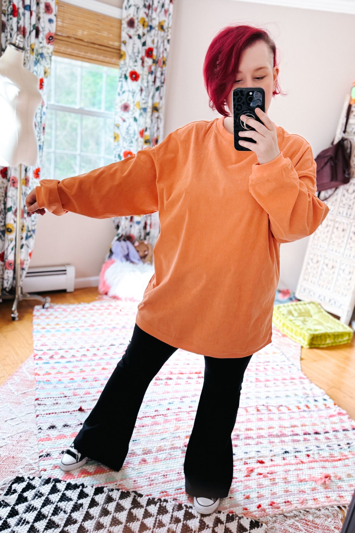 Red haired woman taking a mirror selfie in an oversized orange corded sweatshirt with black flared pants and sneakers in a colorful bedroom