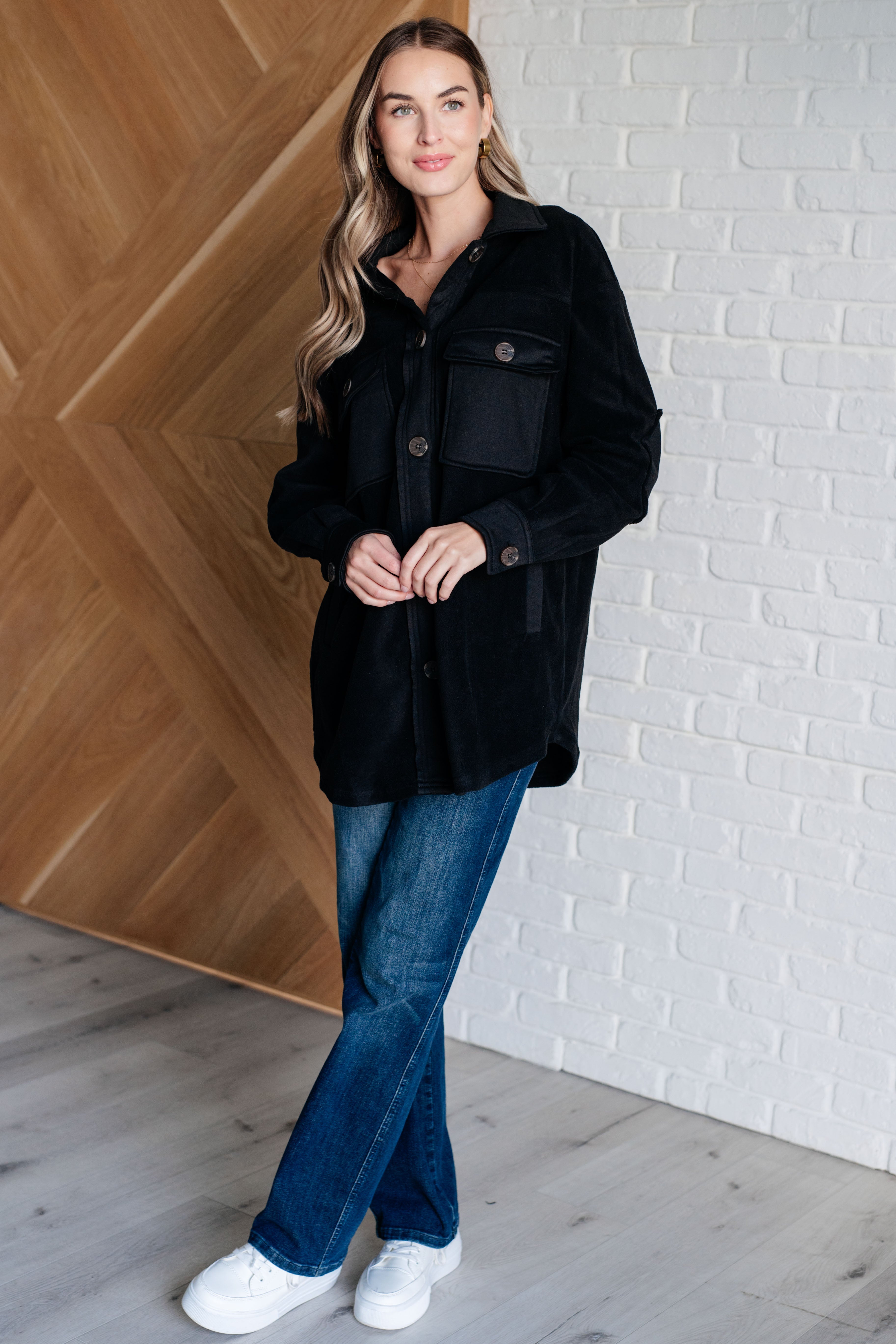 Woman wearing a black fleece button up jacket, blue jeans and white sneakers standing against a white brick wall.