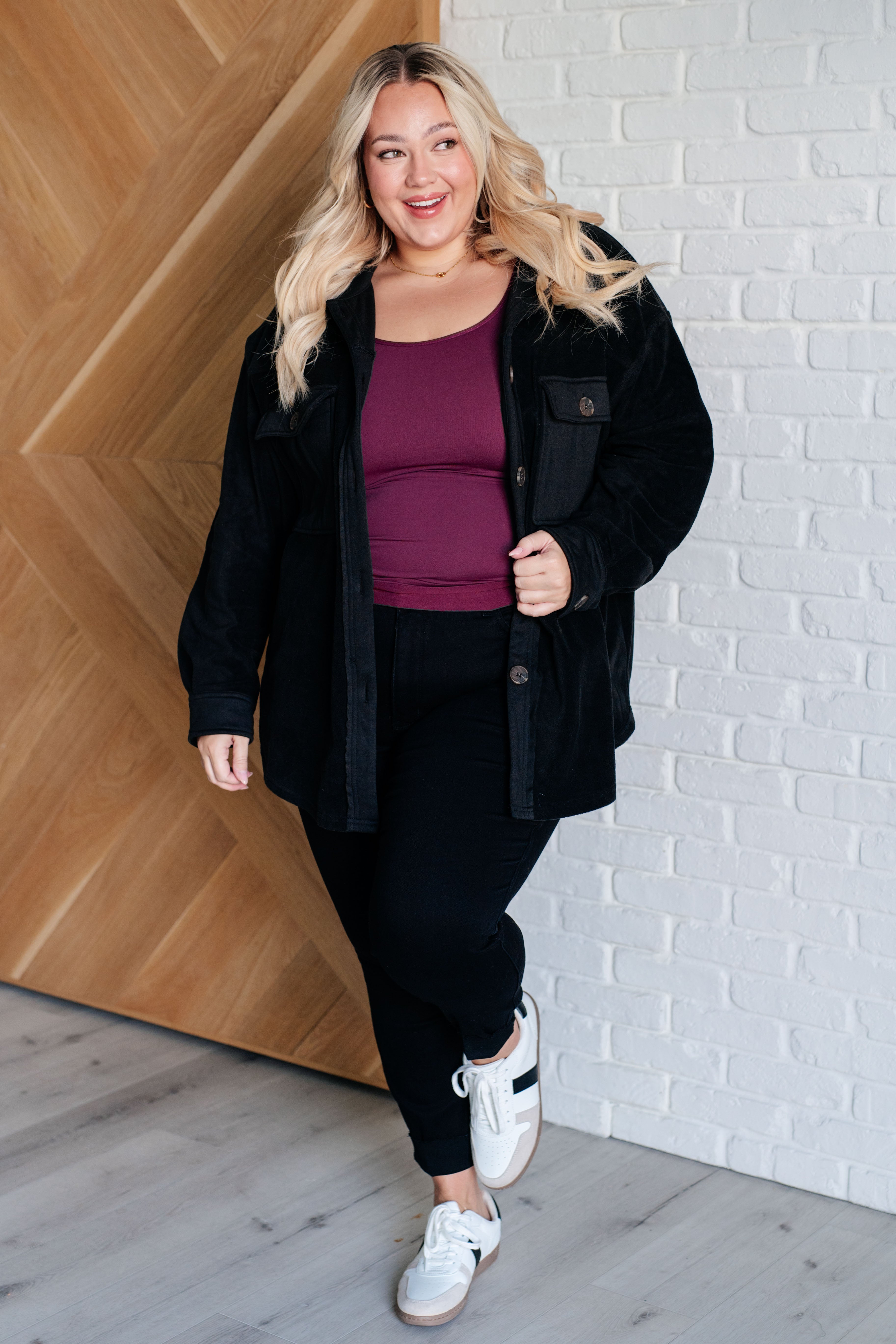 Woman wearing a black fleece button up jacket, a plum top, black pants and white sneakers standing against a white brick wall.