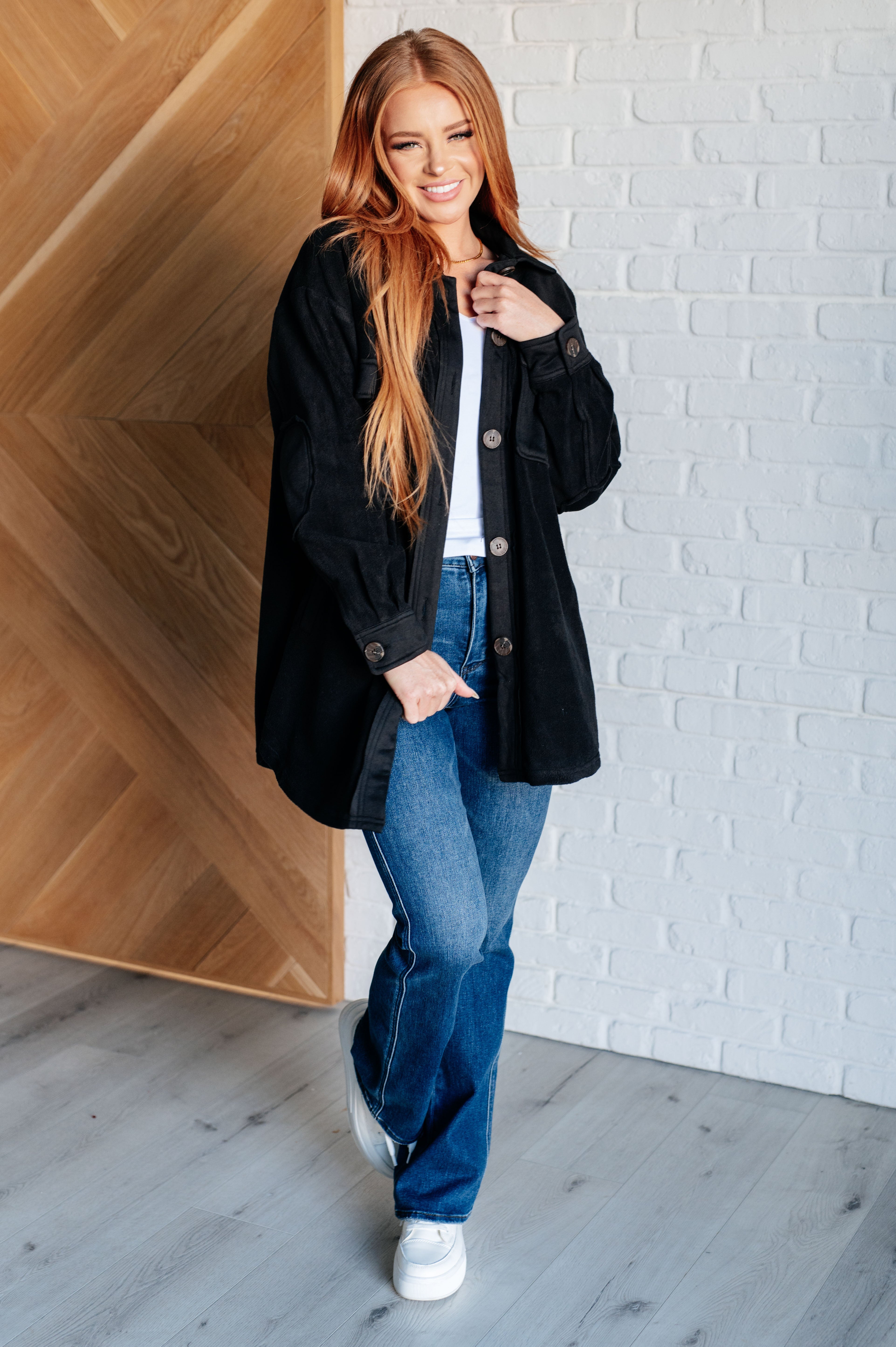 Woman wearing a black fleece button up jacket, a white top, blue jeans and white sneakers standing against a white brick wall.