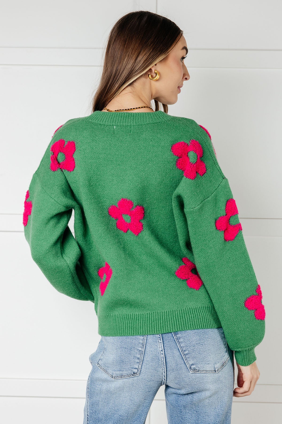 Woman wearing a green sweater embellished with tufted pink flowers and blue jeans against a white wall.
